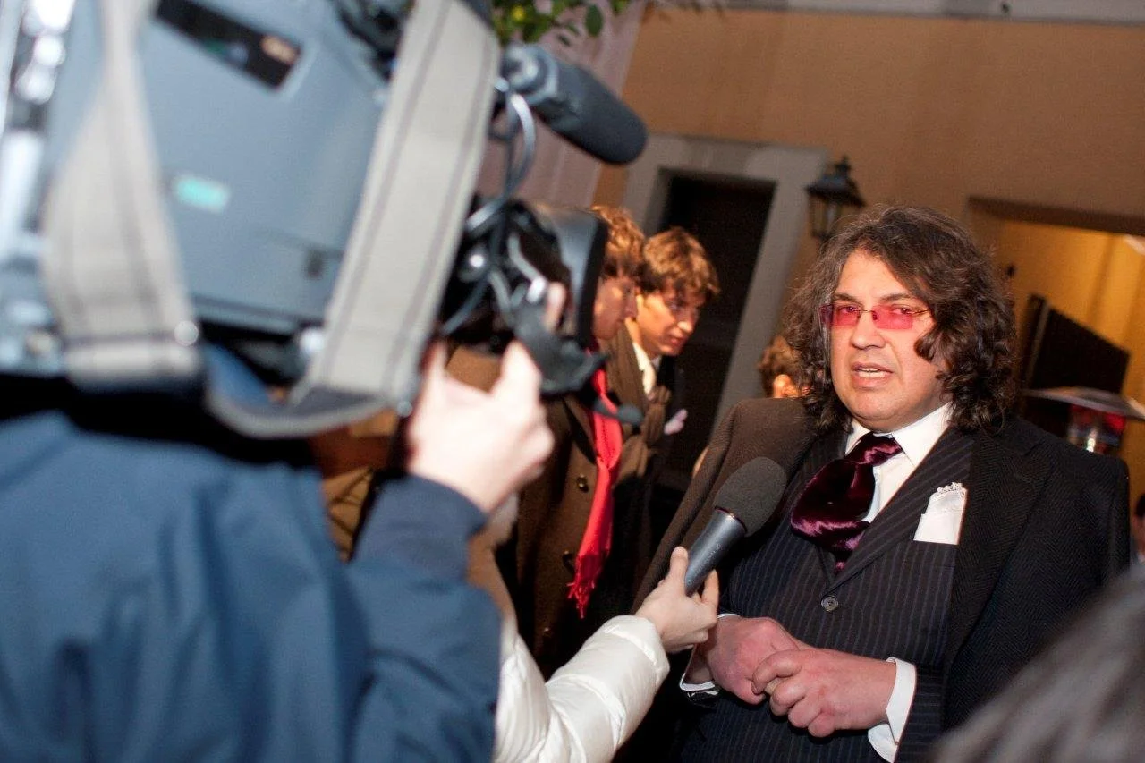 Uomo con capelli ricci e occhiali rosa che parla a una telecamera, circondato da alcune persone.