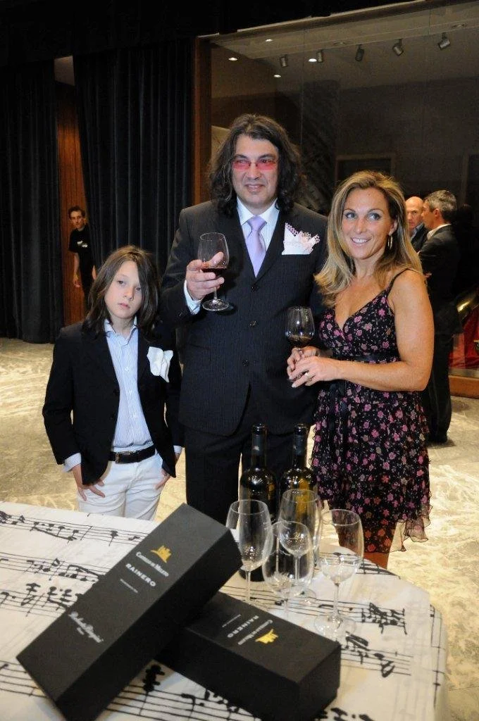 Tre persone sorridenti, un uomo con look alternativo e due donne eleganti, stanno brindando con bicchieri di vino in un ambiente elegante. In primo piano ci sono due bottiglie di vino e altri bicchieri, con una composizione musicale sul tavolo.