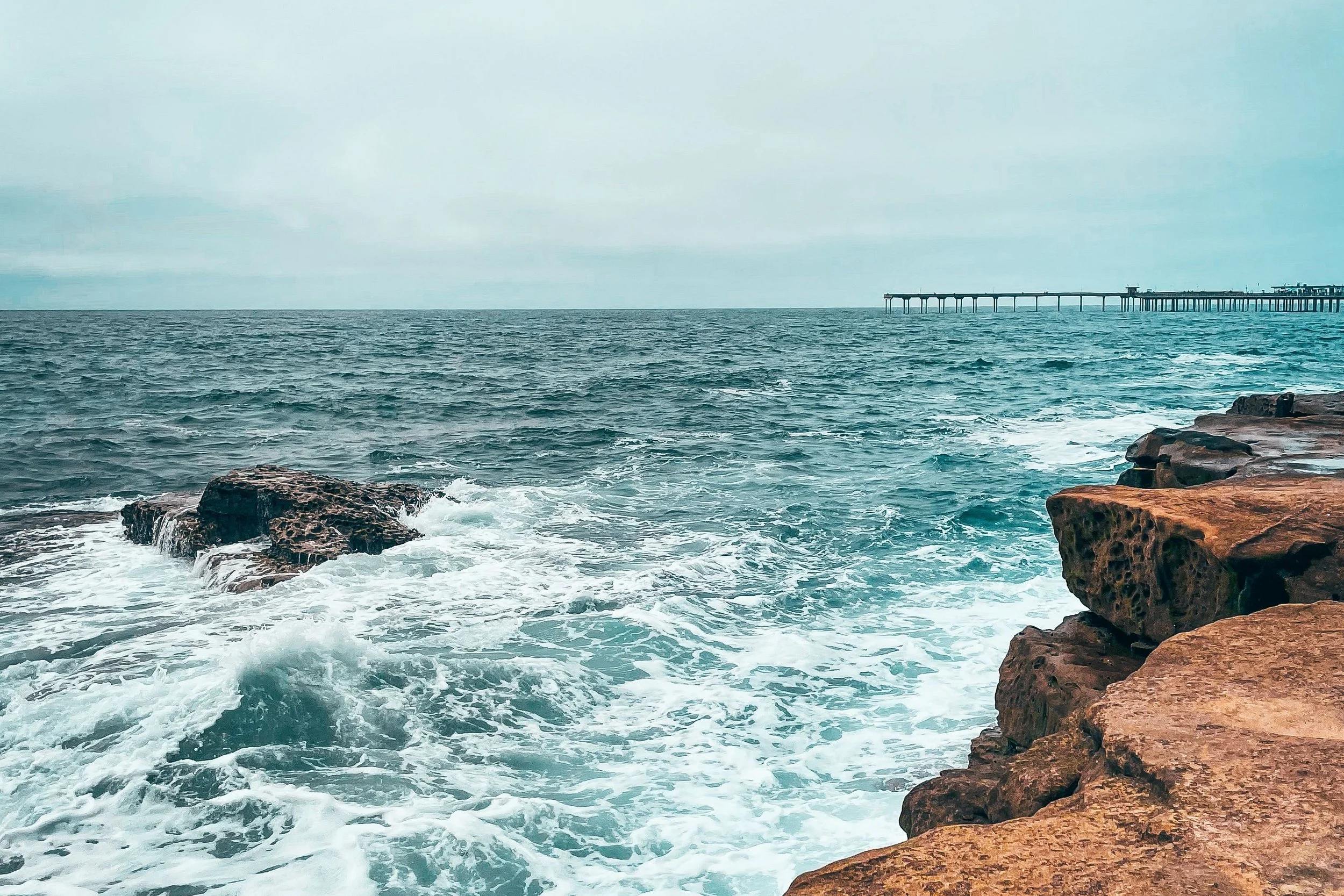 Waves crashing against sandstone rocks along the San Diego coastline, with a long pier stretching toward the horizon under an overcast sky.