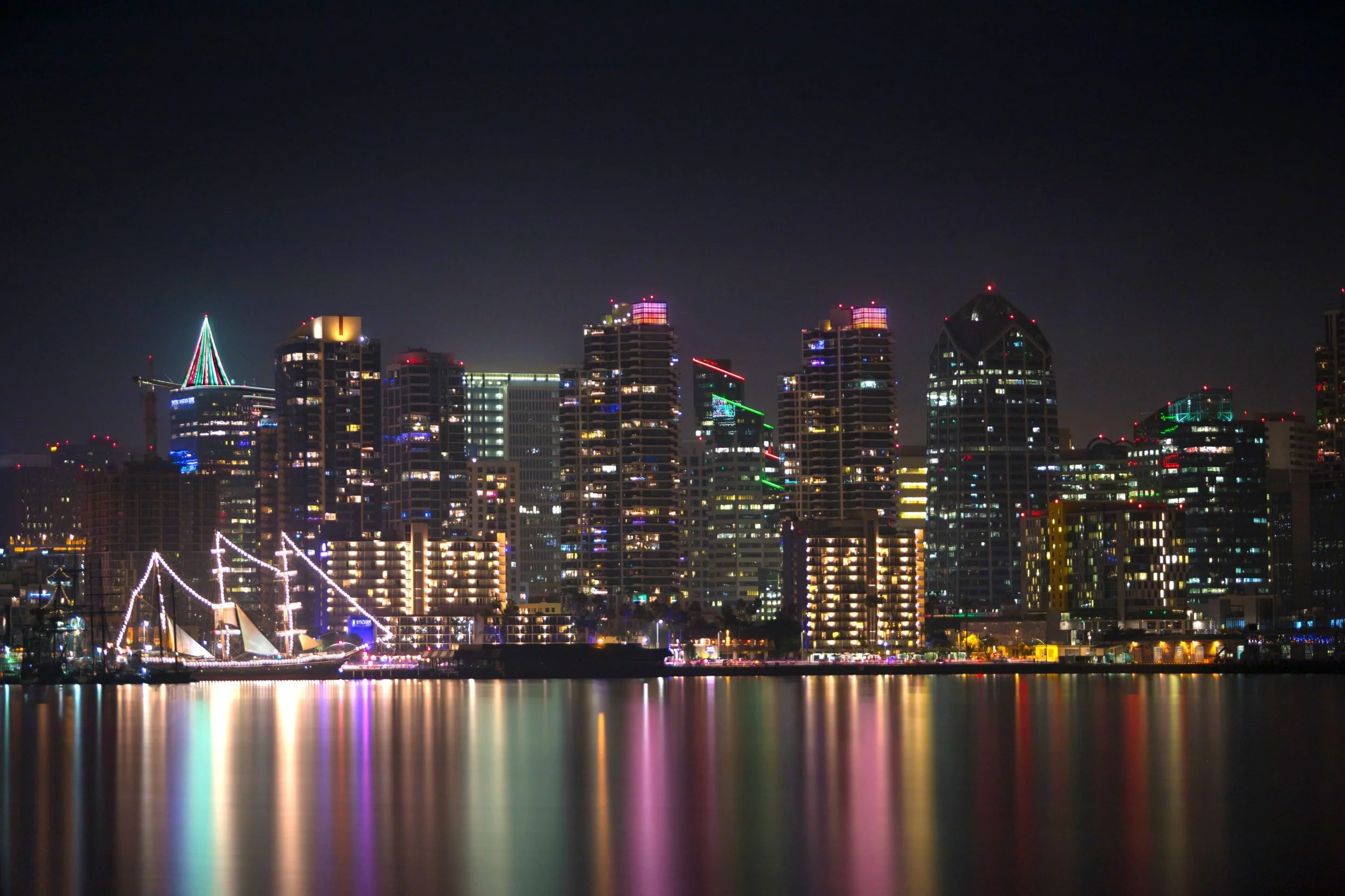 Downtown San Diego skyline lit up at night, with a tall ship strung with white lights and colorful reflections on the bay