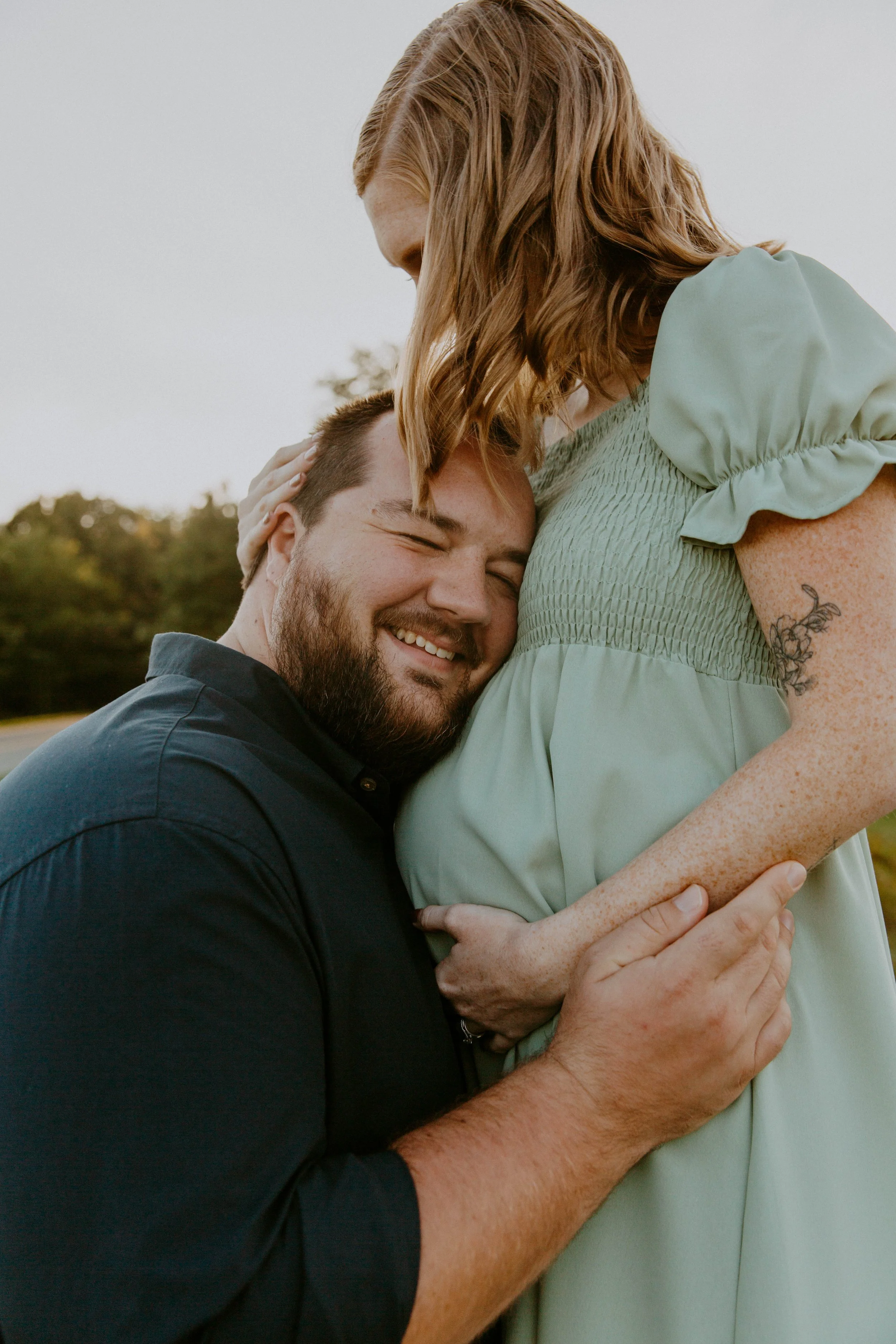 A man and woman share a tender moment outdoors, with the man holding the woman’s pregnant belly and resting his head against it, both smiling.