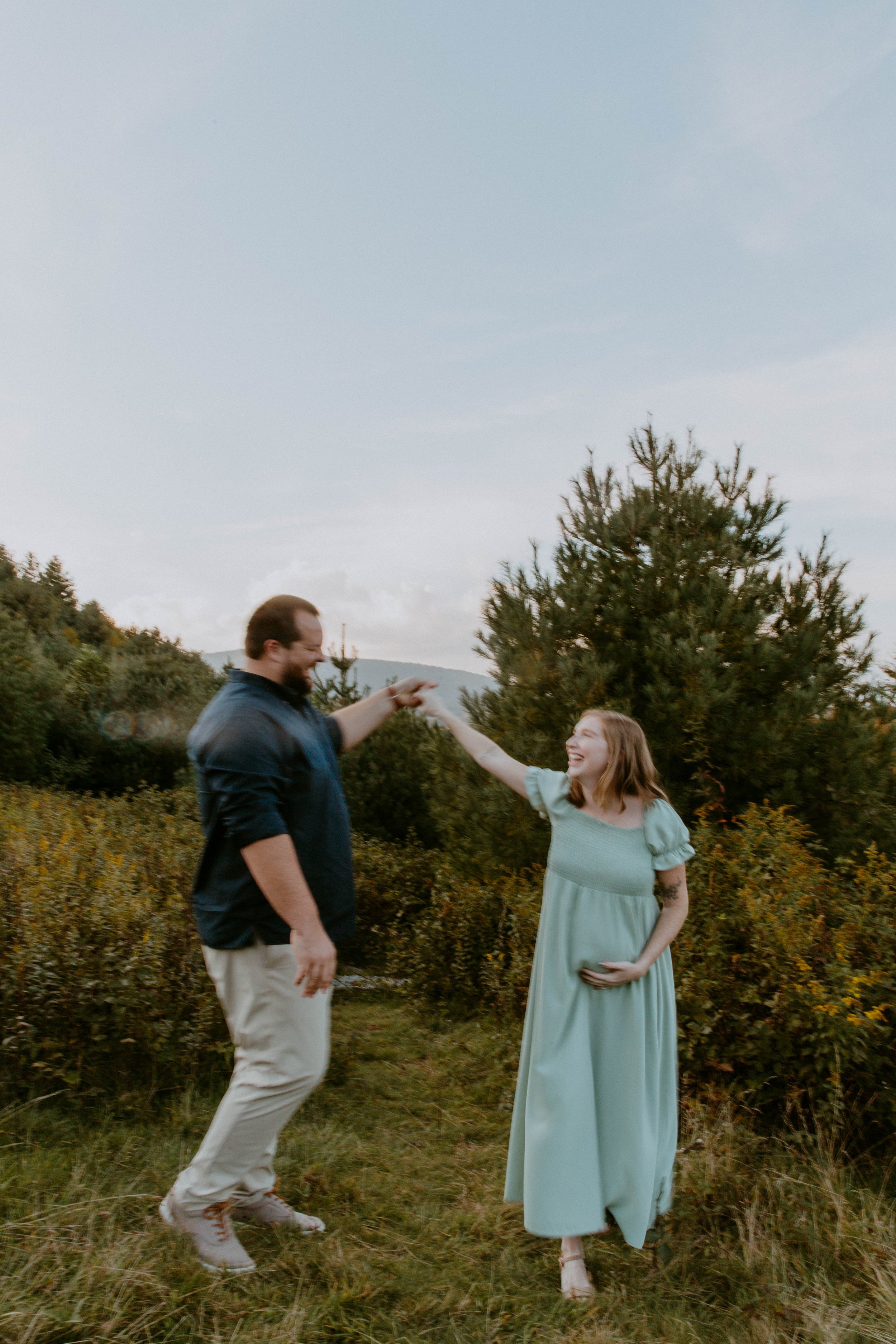 A man and pregnant woman outdoors, smiling and holding hands, with trees and mountains in the background during daytime.