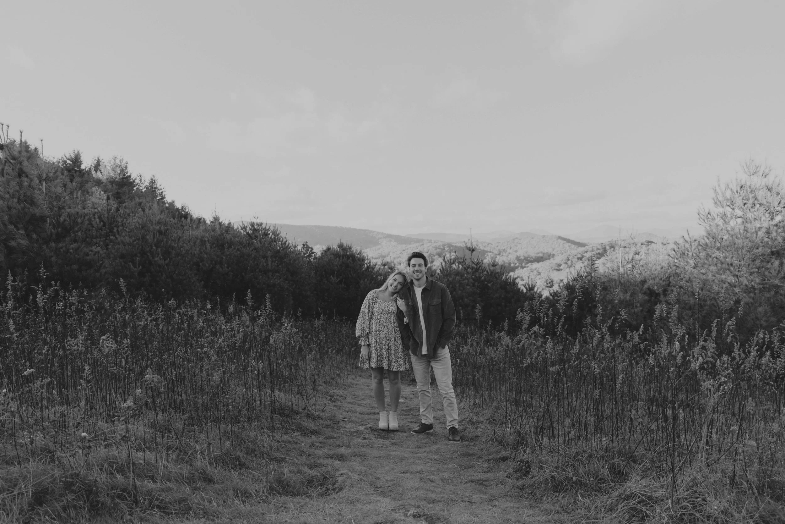 A couple standing on a trail in a grassy field with trees and mountains in the background, smiling on a cloudy day.