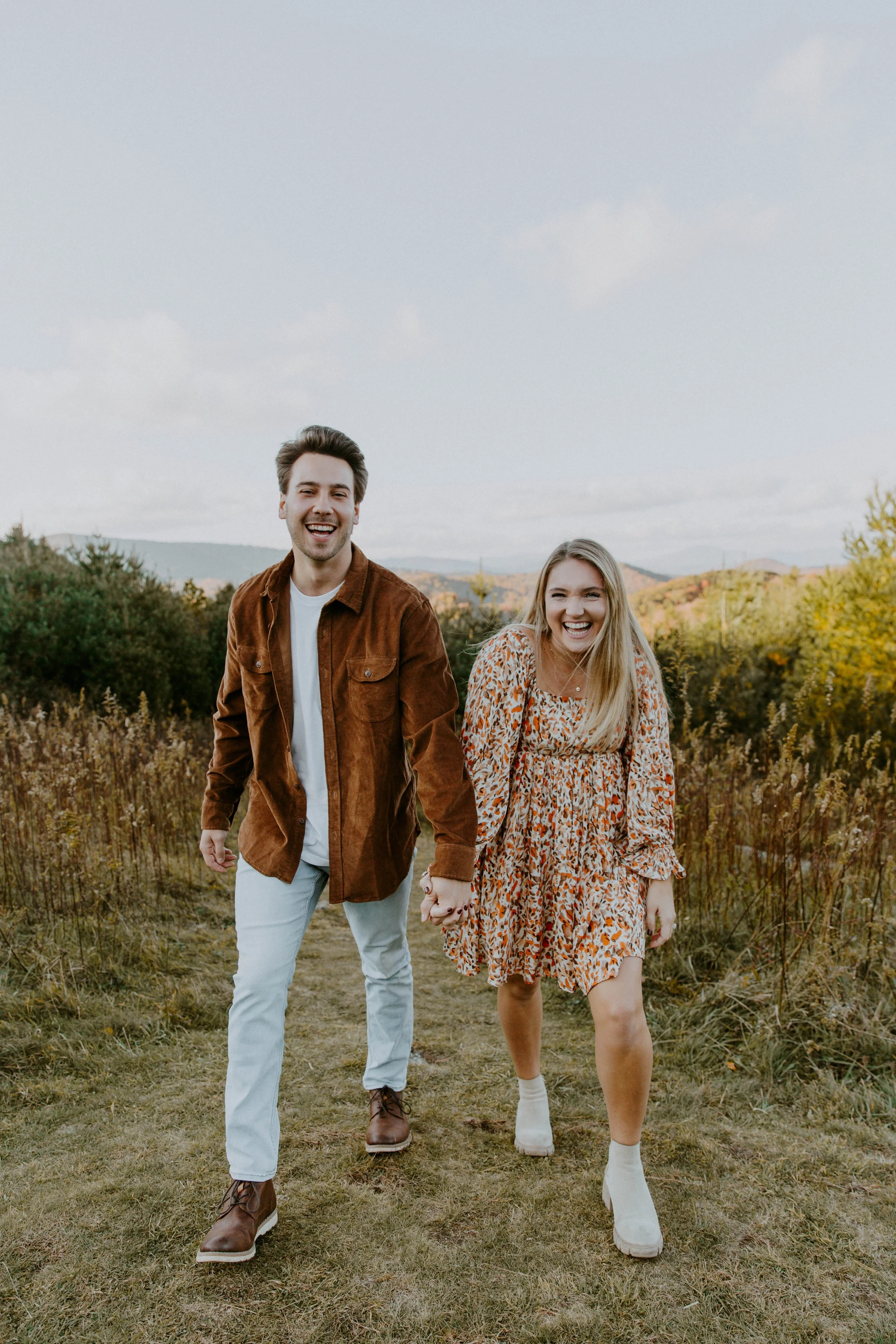 A happy young couple walking hand in hand through a grassy field with autumn foliage and hills in the background.