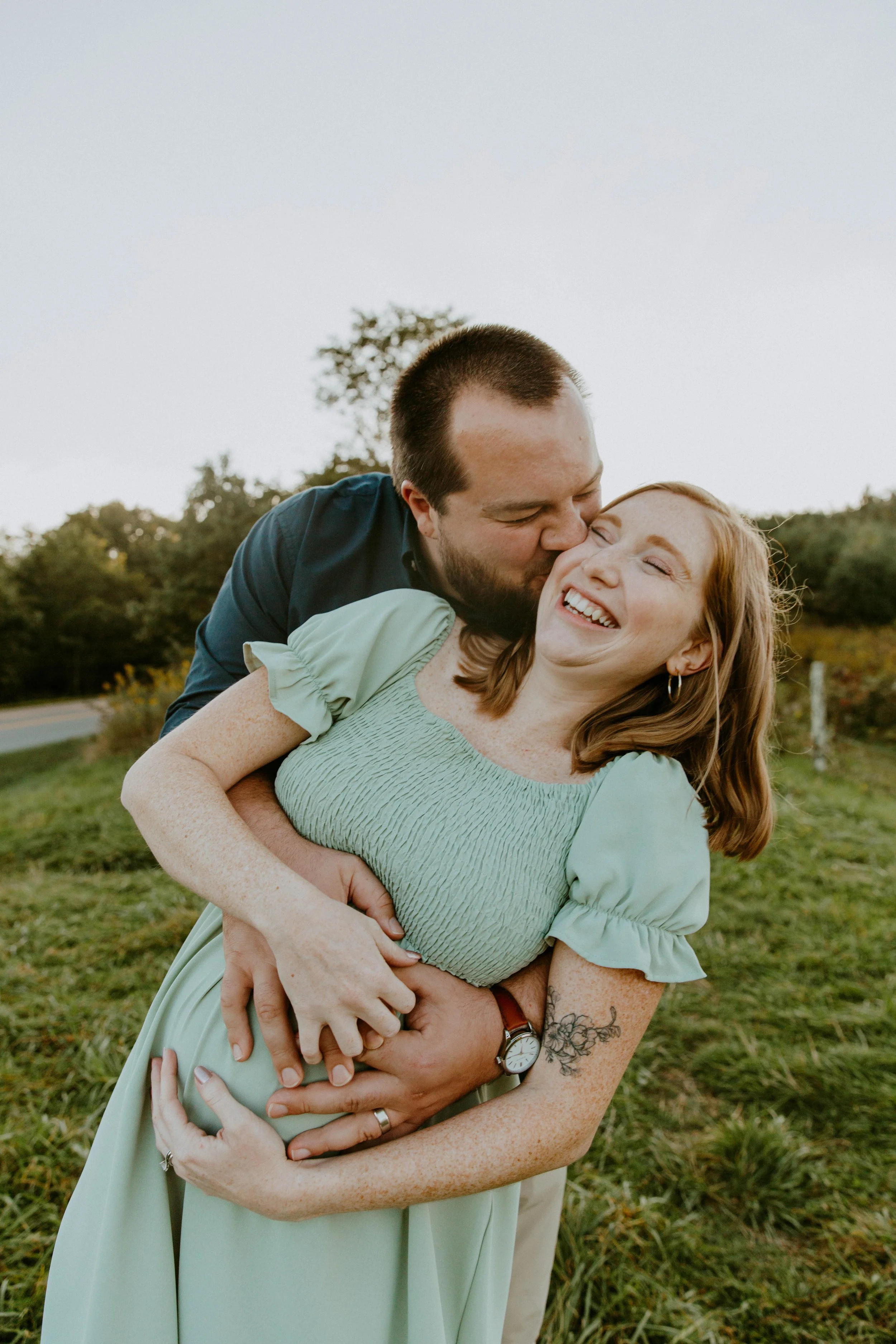 A man is kissing a woman on the cheek while embracing her from behind outdoors, both smiling and showing affection, with greenery and a tree in the background.