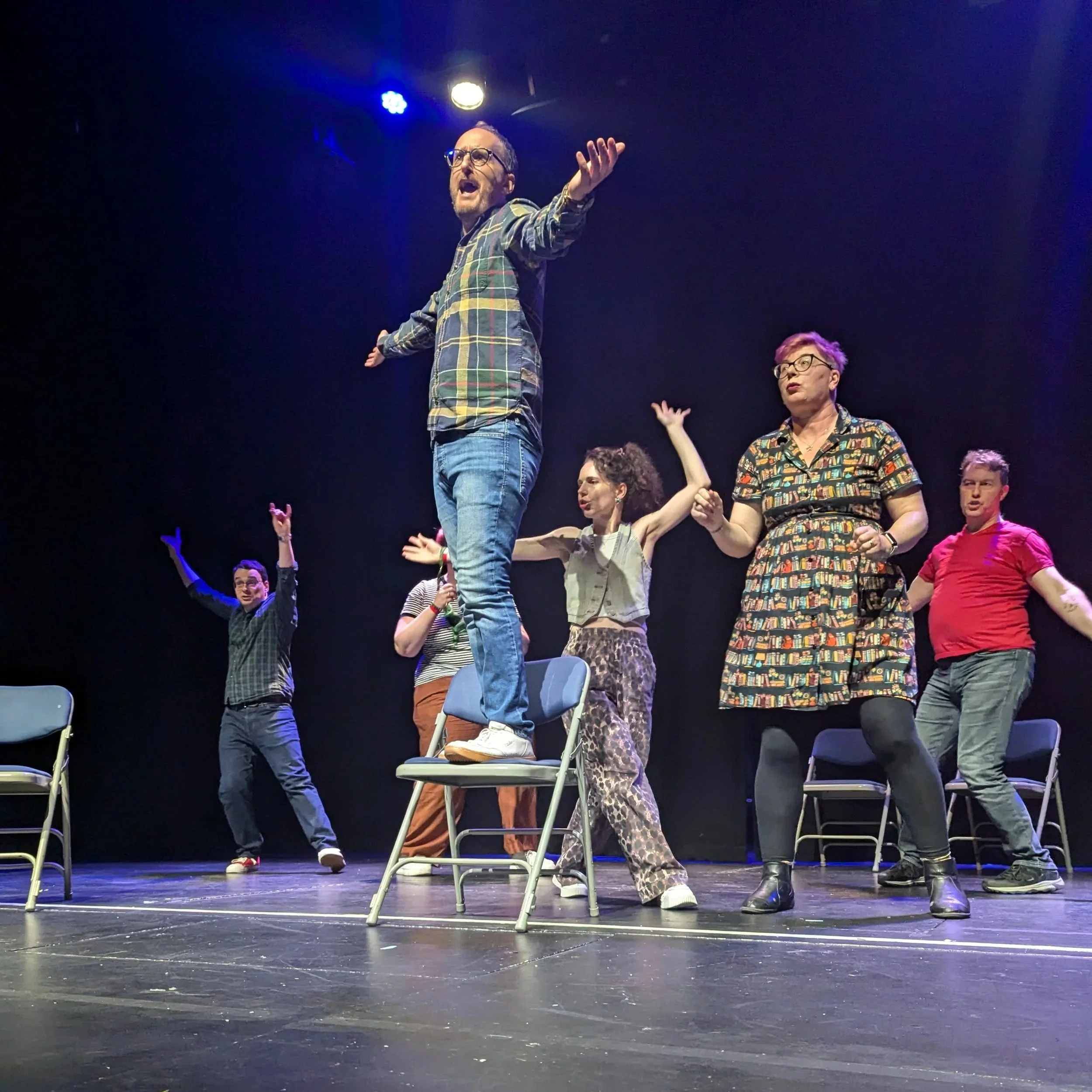 A picture of Rhymes Against Humanity, the improvised musical, with one person standing on a chair, arms outstretched, while surrounded by cast members.
