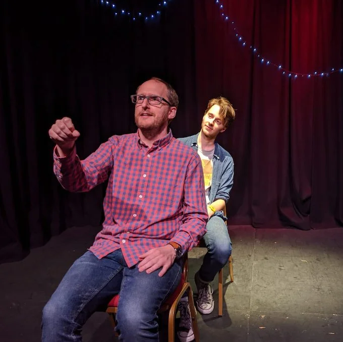 Two improvisers, one sitting in front of the other. The one at the front is miming driving a car. The one behind looks quizzical