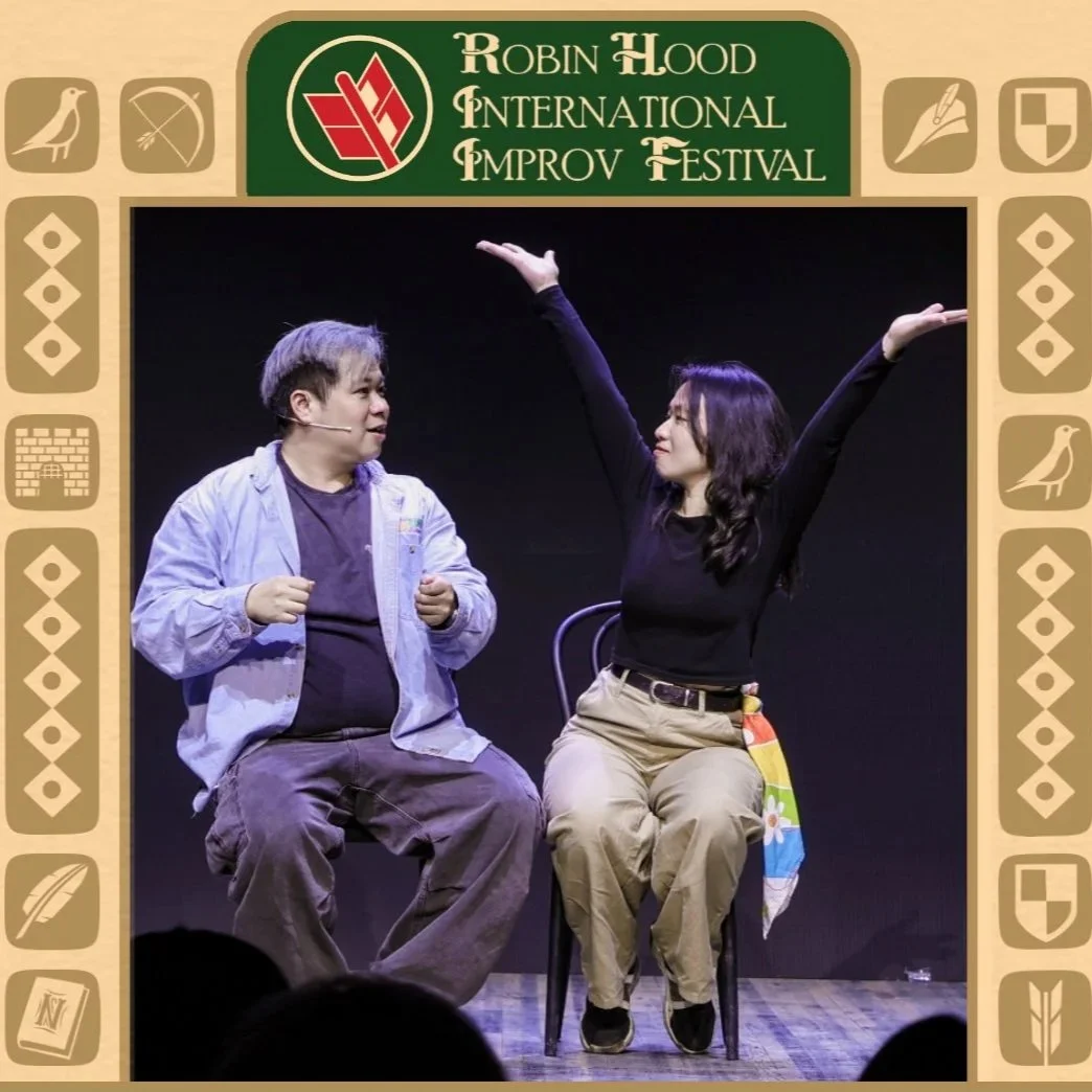 Image shows two improvisers on stage, sitting in chairs. The one on the right has their hands in the air with palms up.