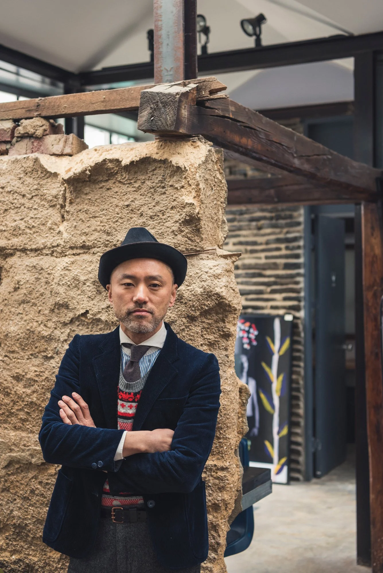 Portrait of a well-dressed Korean man in a jacket and hat leaning against a stone wall in a restaurant
