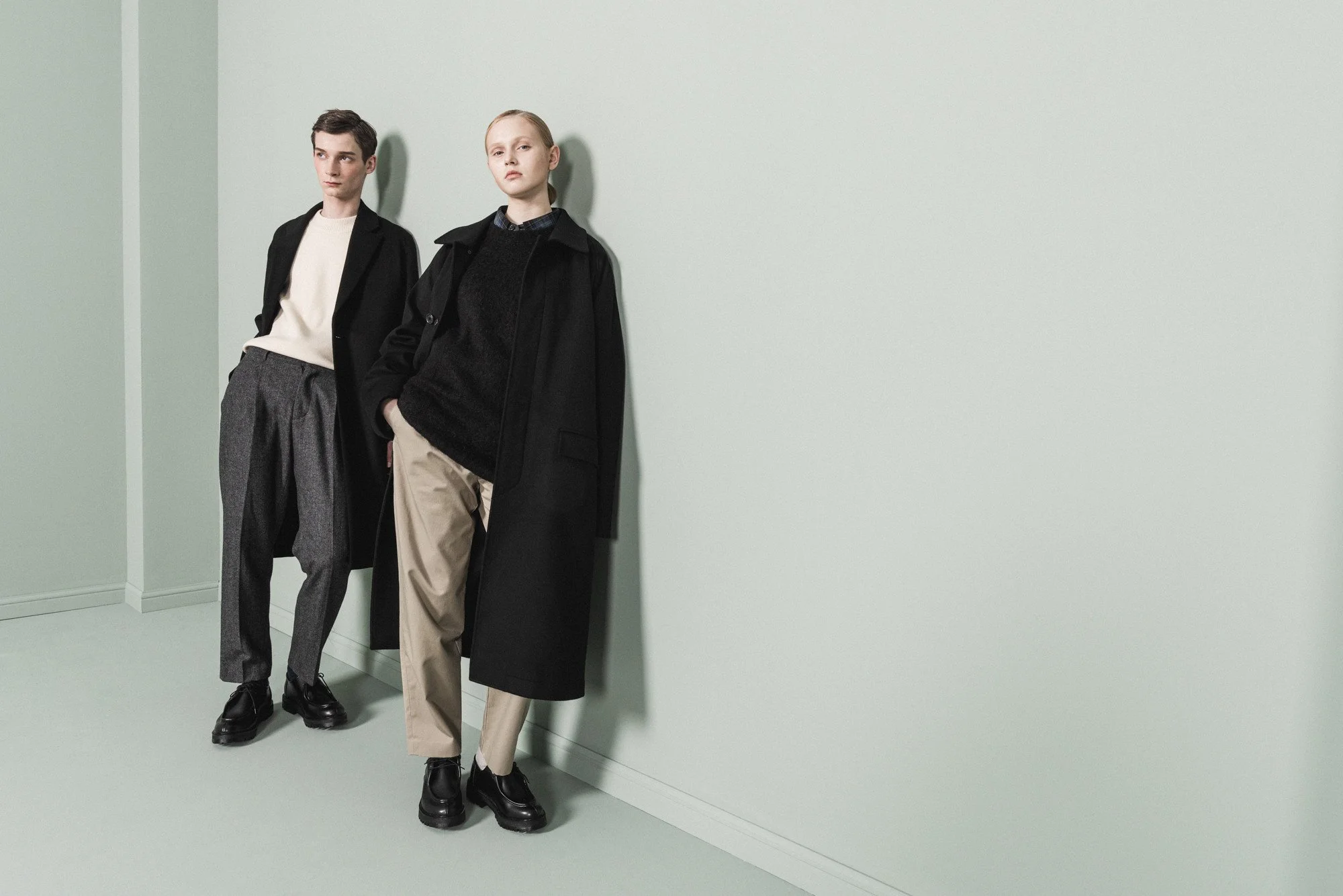 Wide fashion photograph of two models leaning against a wall in a studio