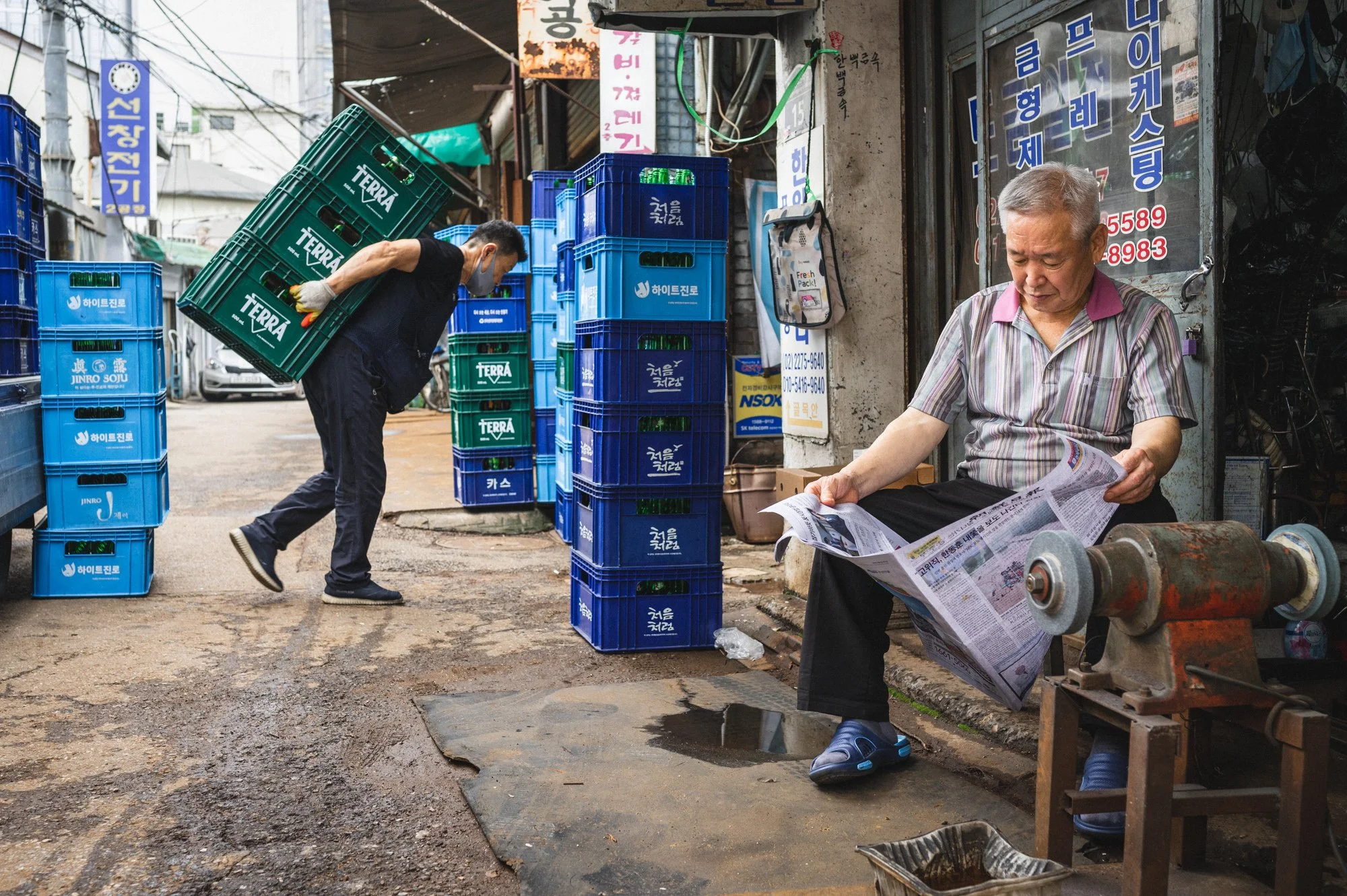 A man reads a newspaper while another carries crates of beer in Euljiro, in Seoul, South Korea