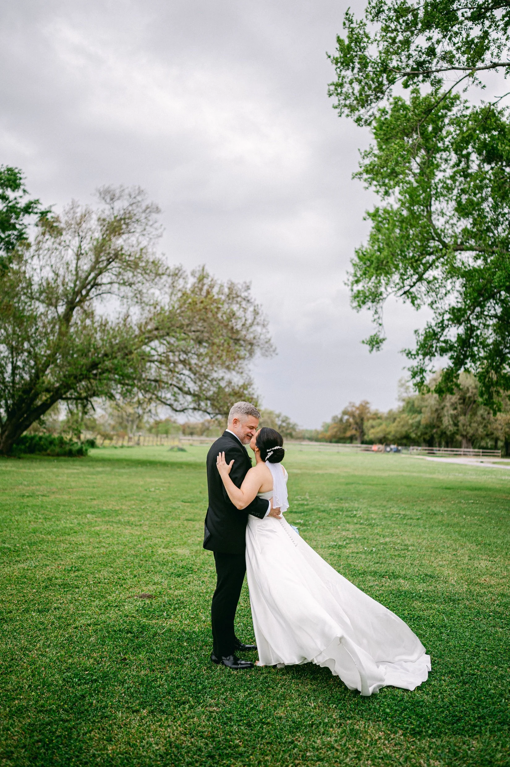 A bride and groom dancing outdoors on green grass, surrounded by trees with overcast sky.