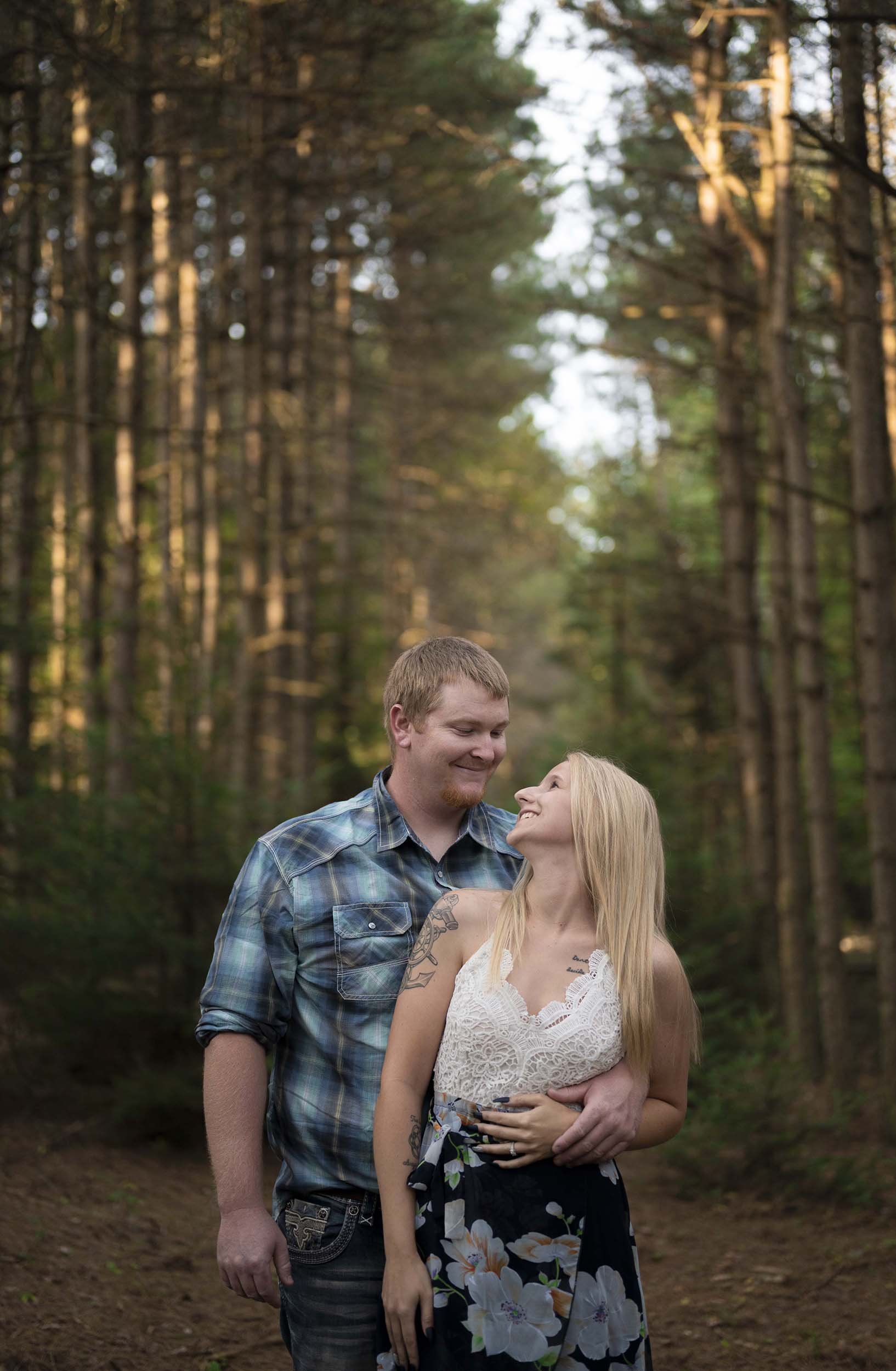 A smiling couple standing closely together in a forest with tall trees, sunlight filtering through, and foliage in the background.