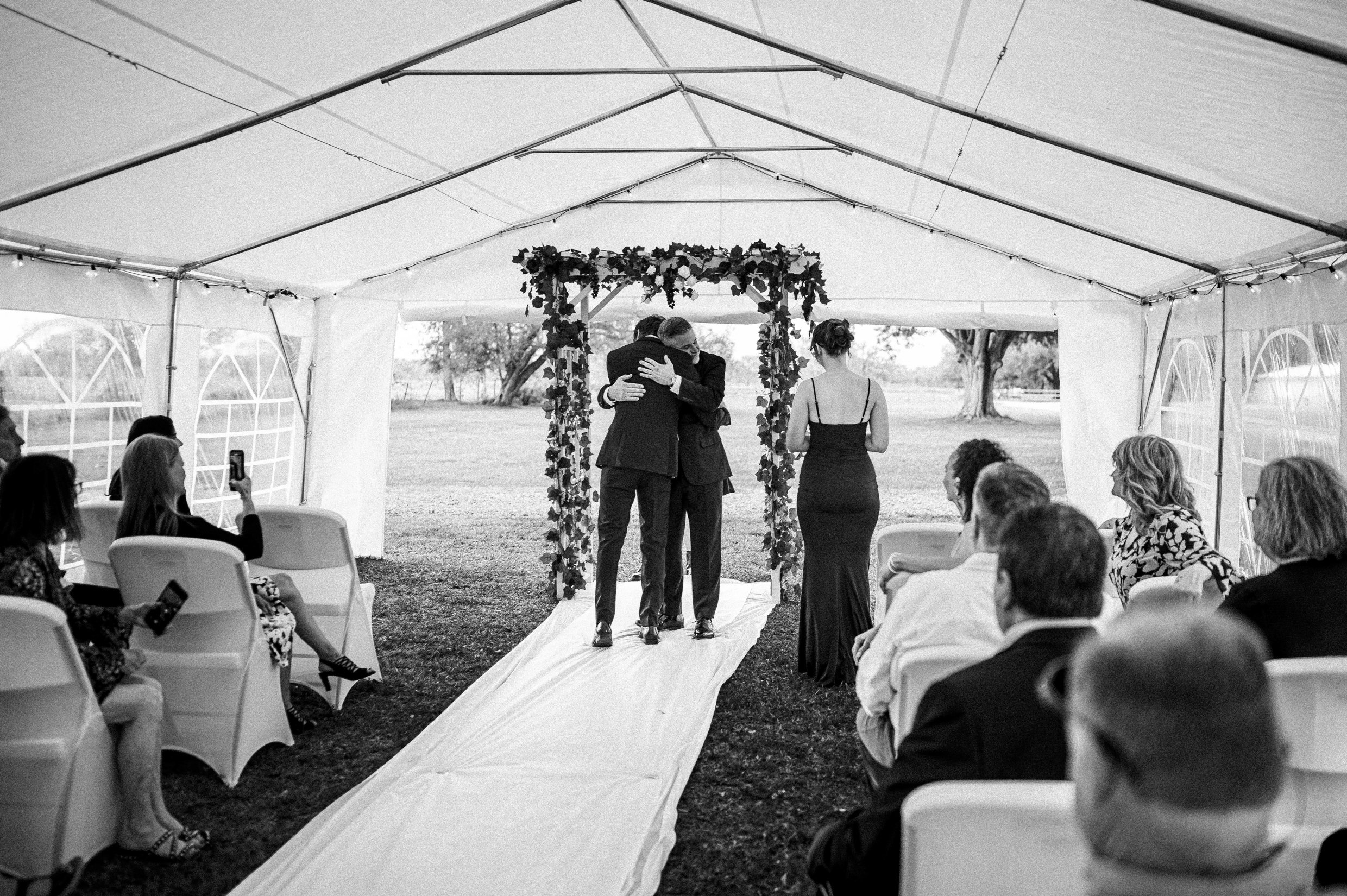 A groom hugs his children during a wedding ceremony taking place inside a tent outdoors.