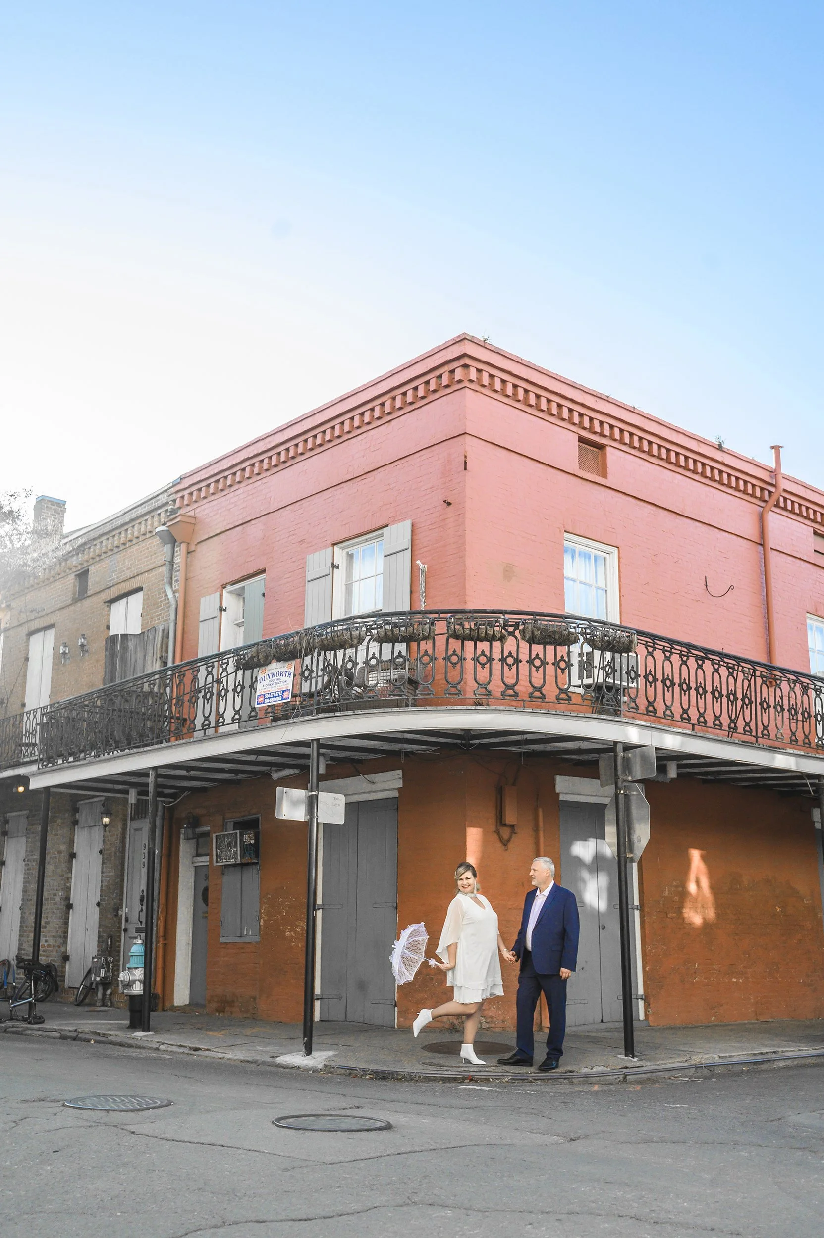 A newlywed couple holding hands and smiling at each other on a New Orleans French Quarter street corner with a pink brick building and wrought-iron balconies.