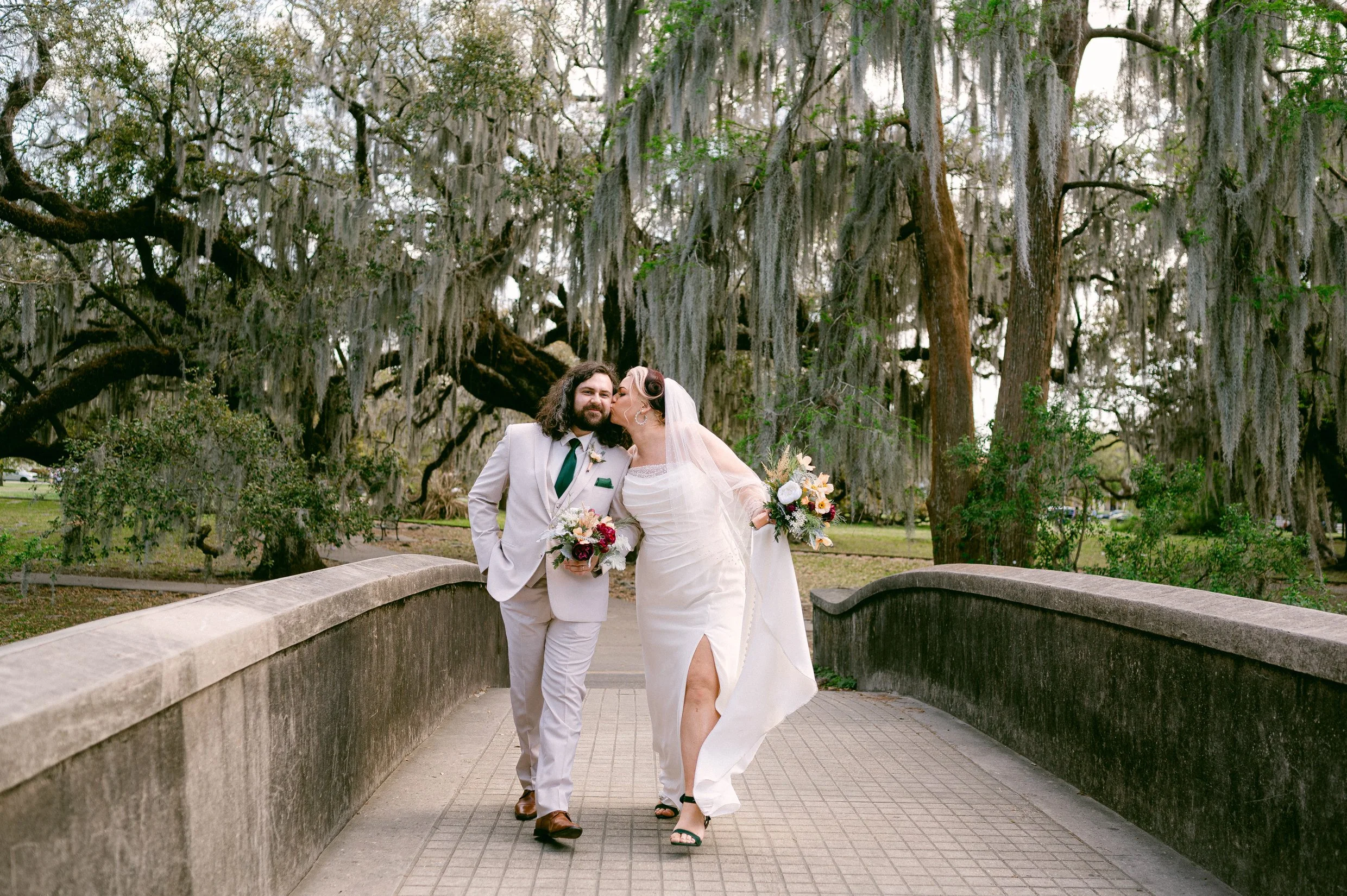 A newlywed couple walking on a stone bridge in New Orleans City Park, surrounded by large moss-covered oak trees.