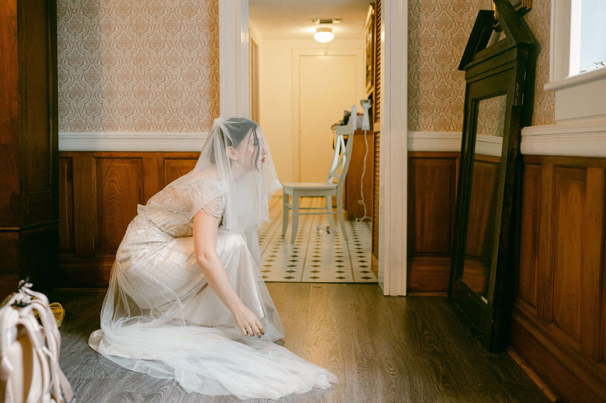 A bride in a wedding dress with veil sitting on the floor in a hallway, with mirror leaning against the wall.