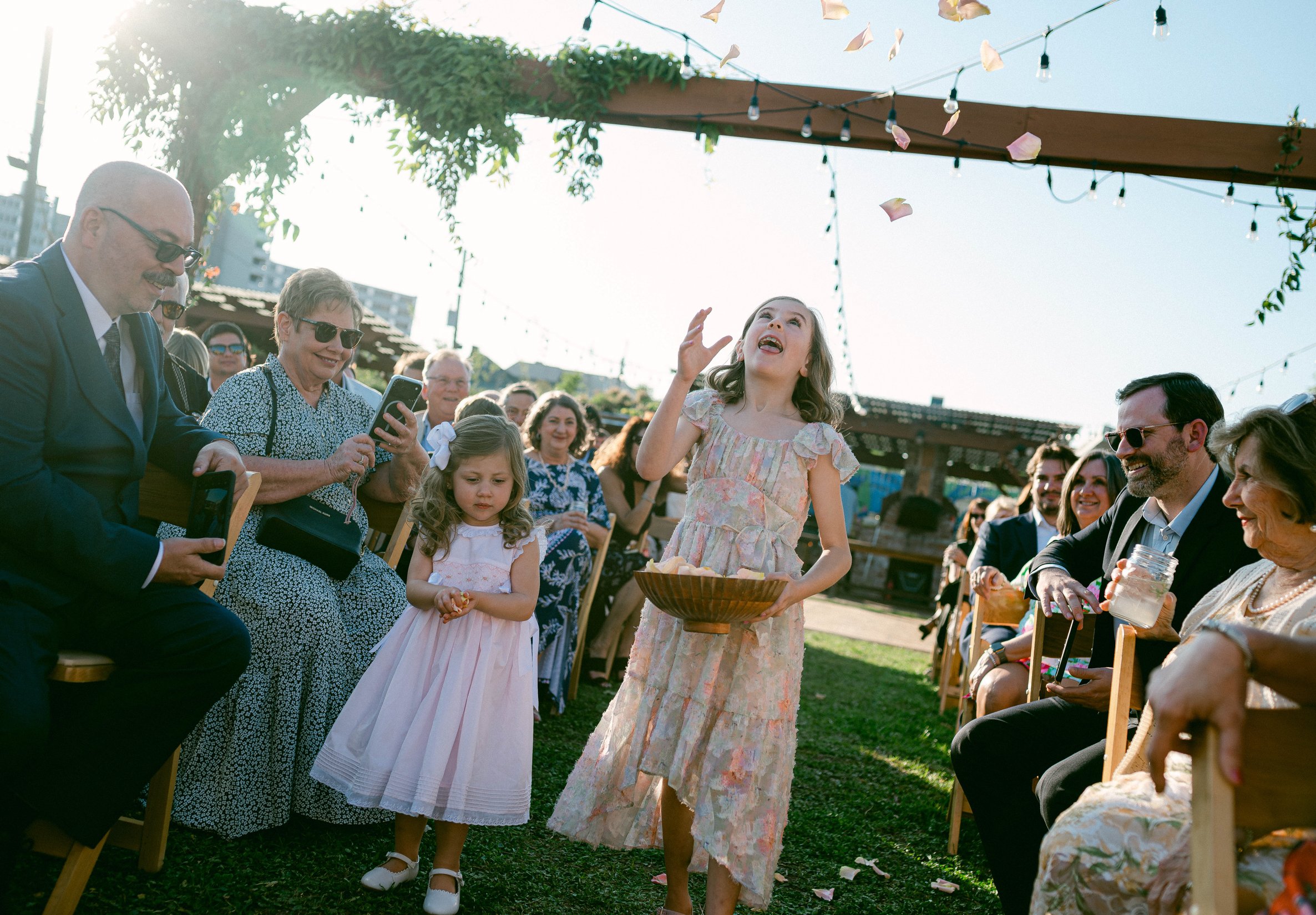 A flower girl tossing flower petals into the air at an outdoor wedding, surrounded by happy guests.