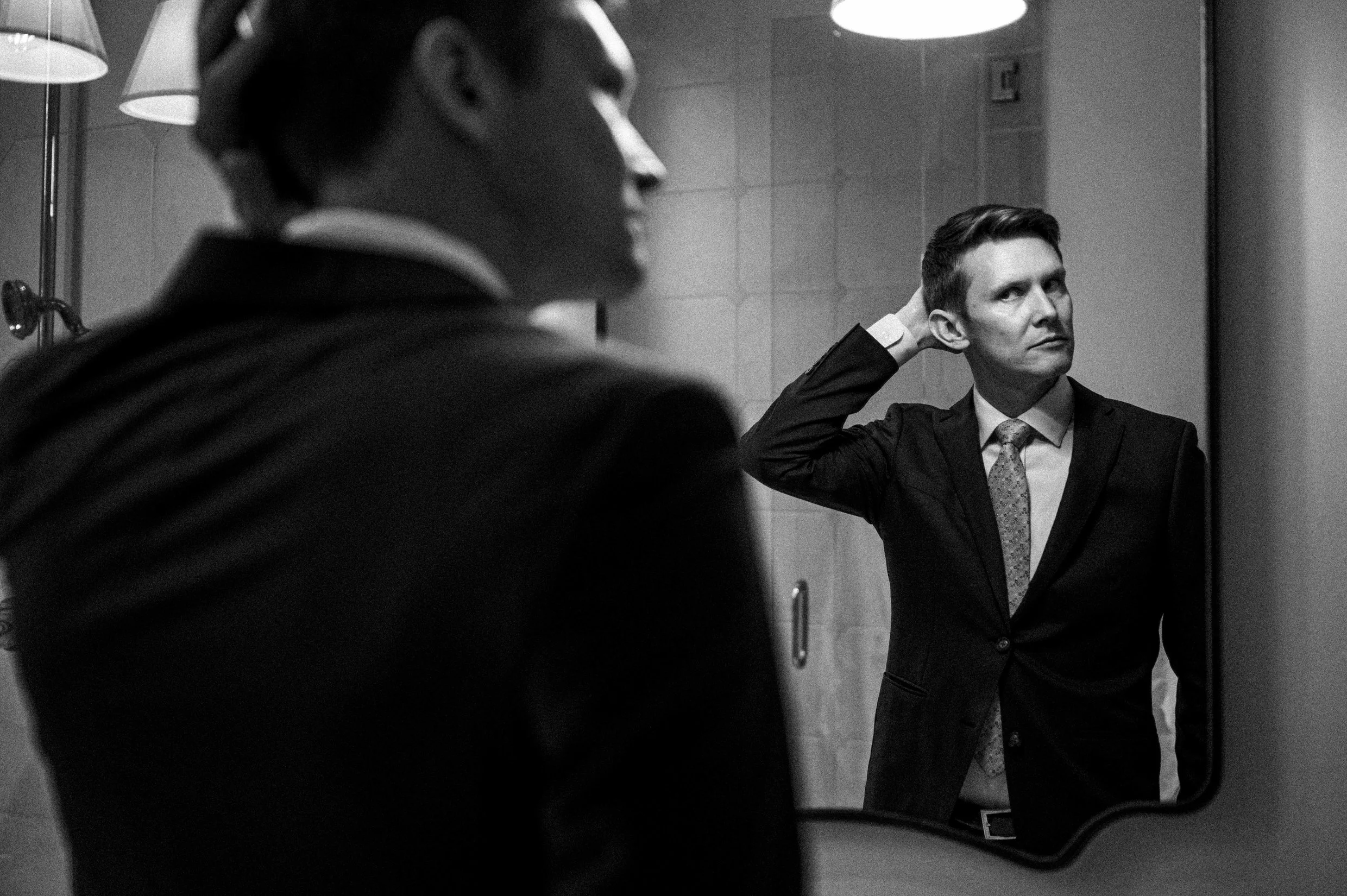 A groom in a suit looking at his reflection in a bathroom mirror.