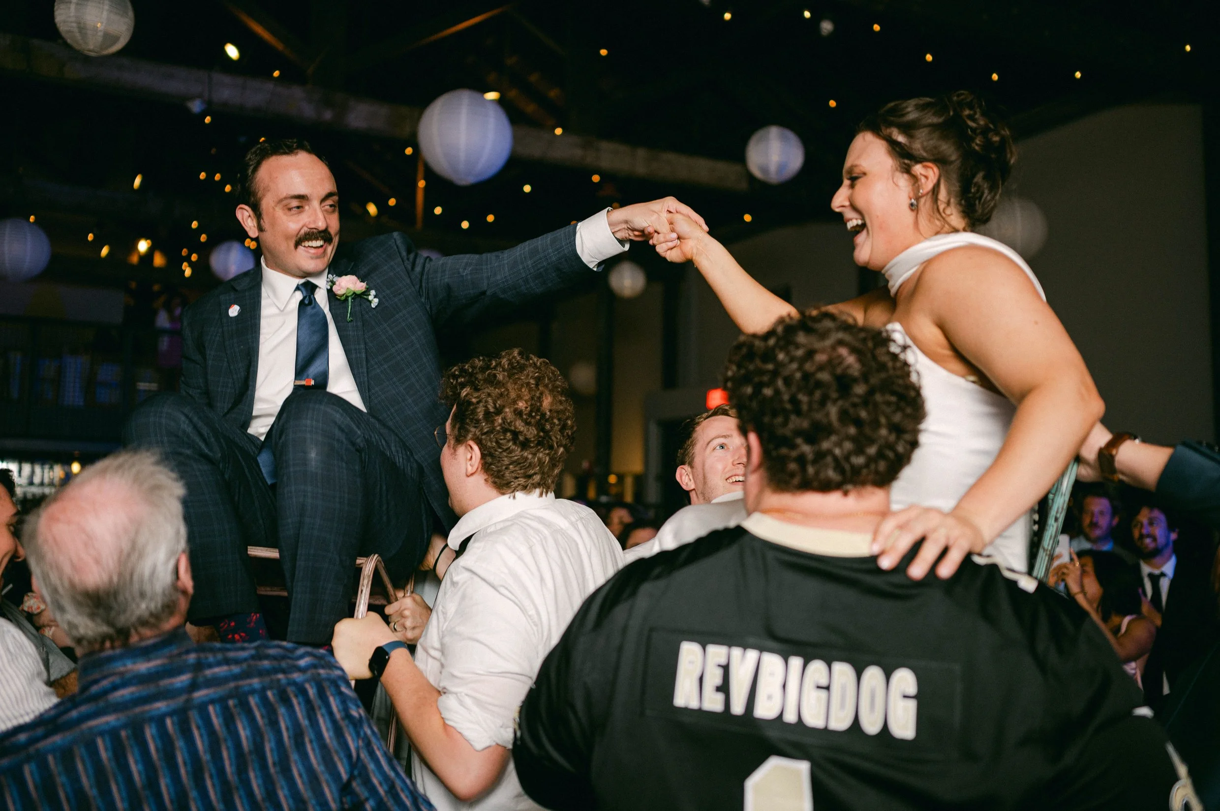 People celebrating at a wedding reception, with one woman being lifted on a man's shoulders, both smiling and laughing.