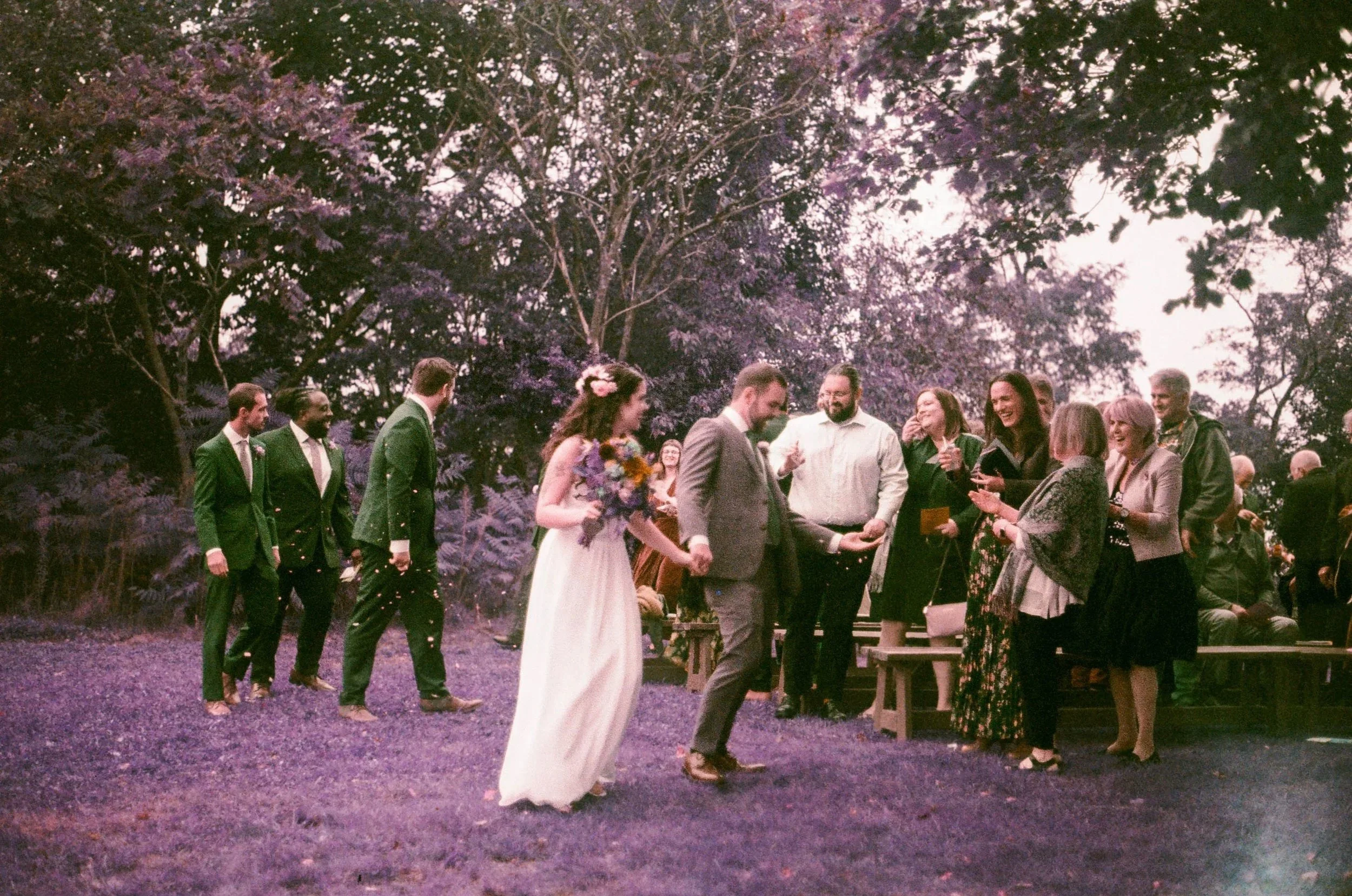 The crowd at an outdoor wedding ceremony, with the bride and groom at the center holding hands, surrounded by friends and family, under a large purple-flowered tree during dusk.