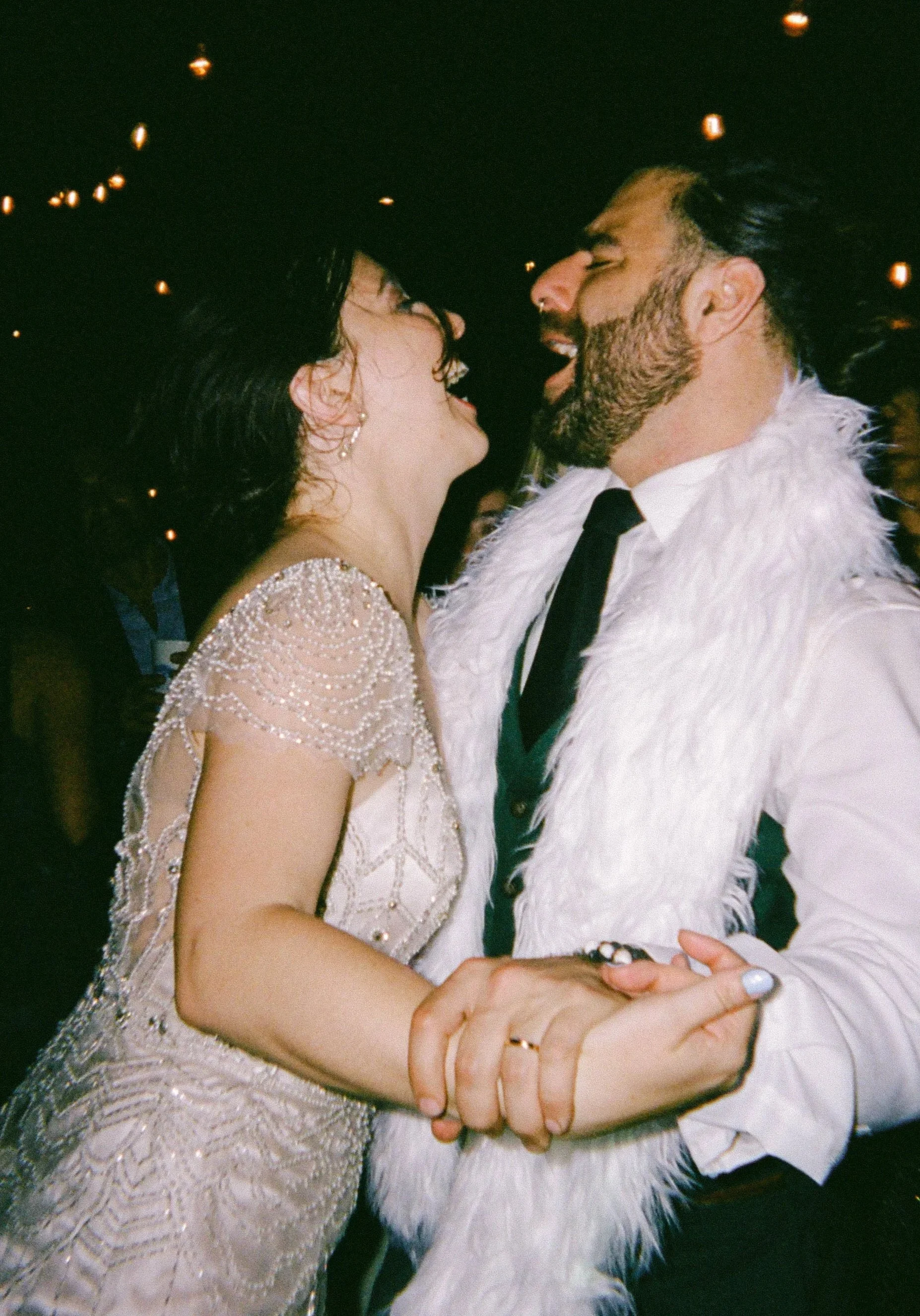 A woman and a man are dancing closely at their wedding, smiling and holding hands, with dark background and festive lights.