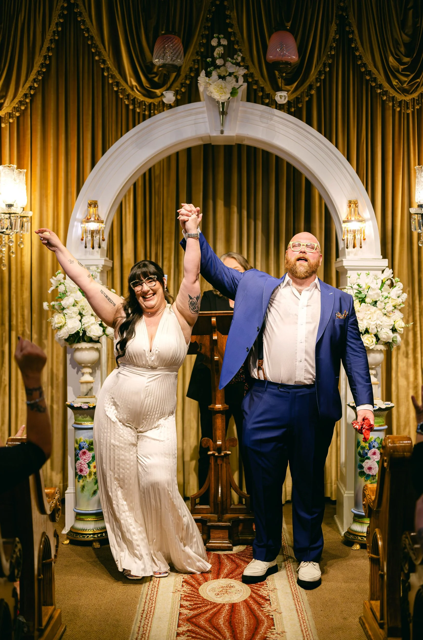 A couple at their wedding ceremony, holding hands and raising them in celebration, standing under a decorated arch with floral arrangements in a New Orleans wedding chapel.