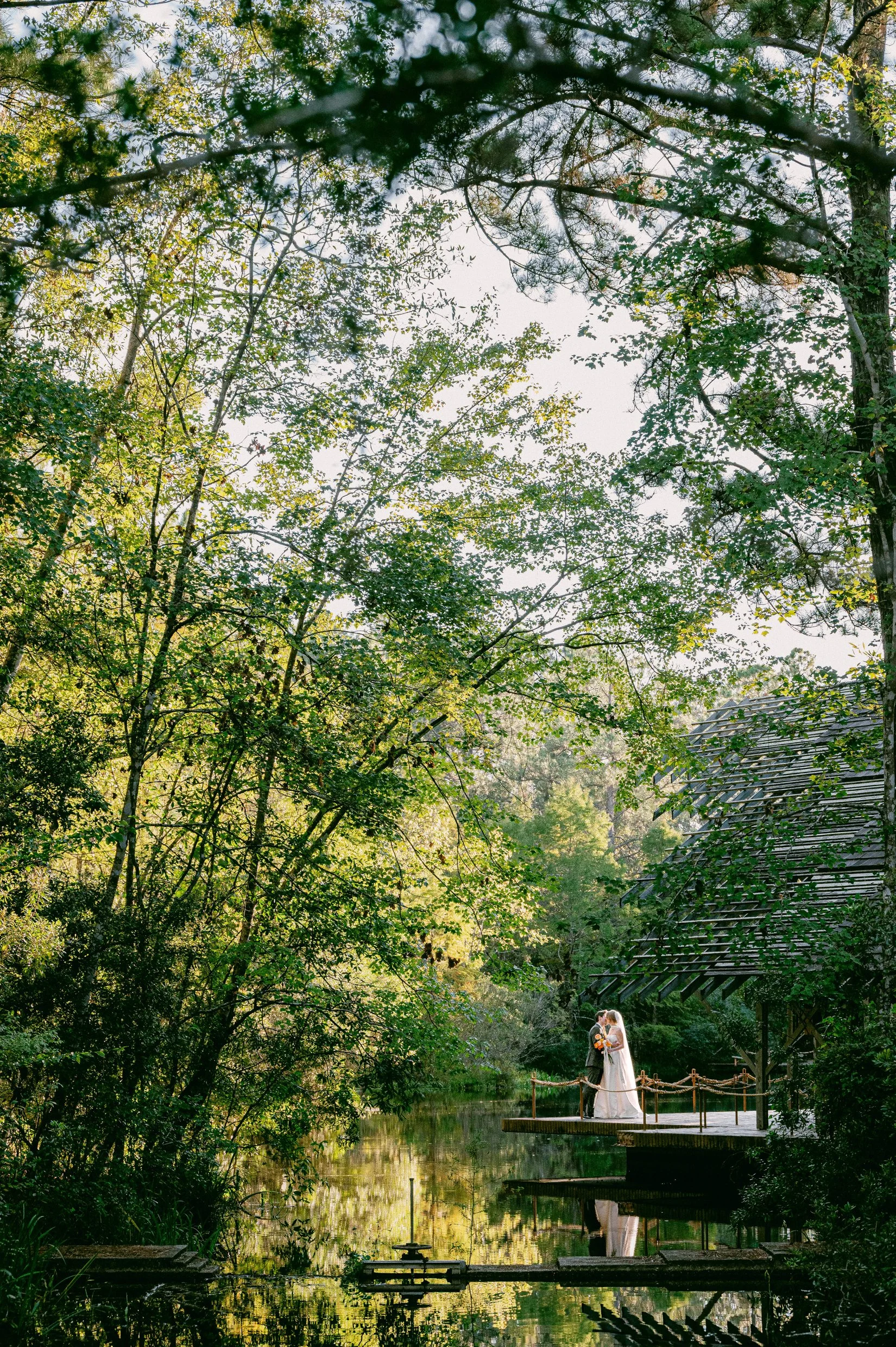 A bride and groom standing on a small dock by a pond, surrounded by lush green trees and foliage, with sunlight filtering through the leaves, at Crosby Arboretum in Mississippi.
