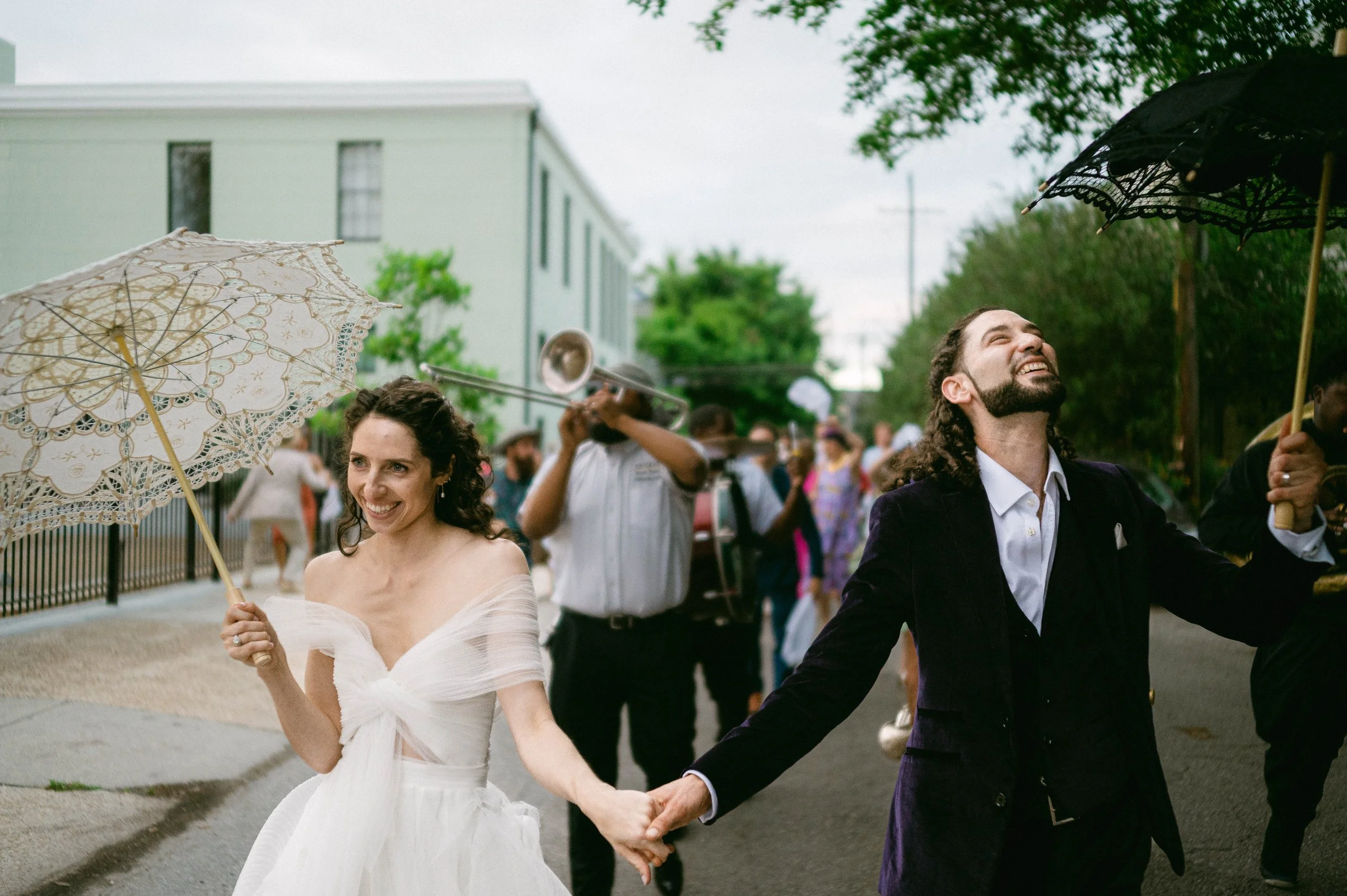 A joyful bride and groom holding hands and smiling at each other, with a crowd and marching band in the background, during an New Orleans wedding second line.