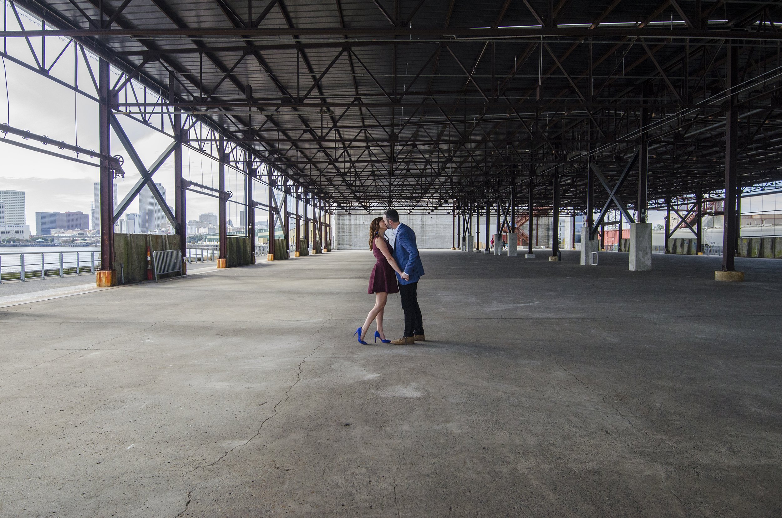 An engaged couple sharing a kiss under an industrial structure by the waterfront with the New Orleans skyline in the background.
