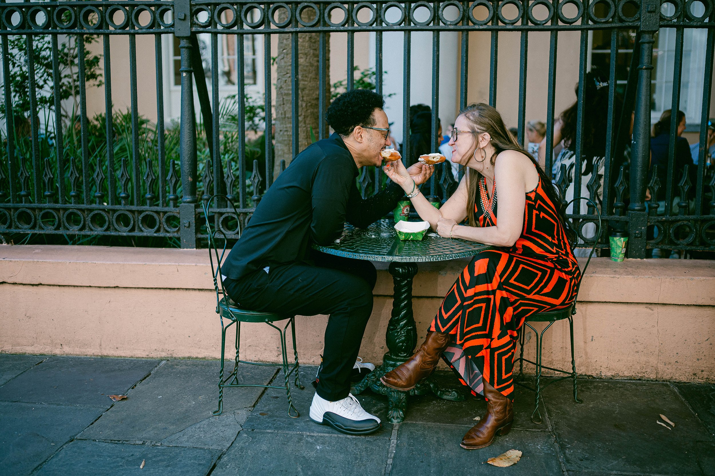 A man and woman sharing beignets at an outdoor cafe table behind a black wrought iron fence in New Orleans.