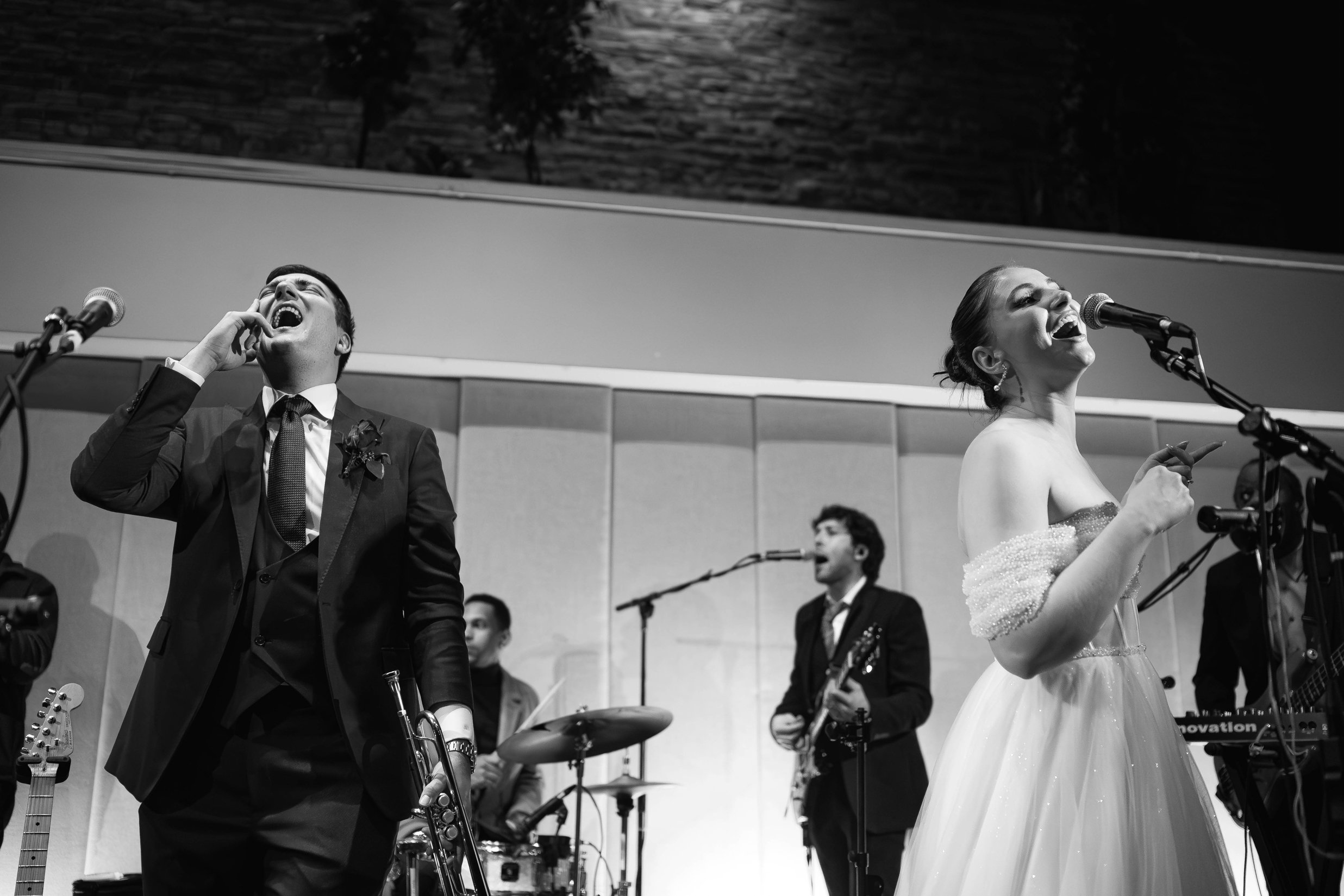 Black and white photo of a bride and groom singing into microphones on stage at their wedding, other performers in the background.