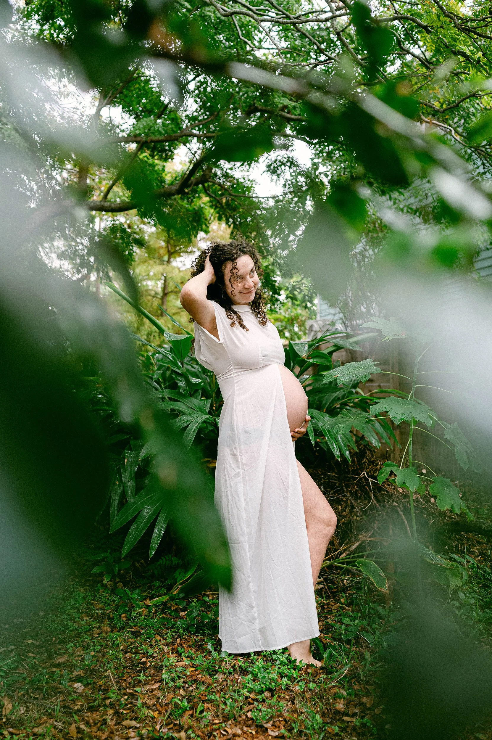 A pregnant woman in a white dress standing in a lush, green garden surrounded by various plants and trees, with a smile and her hands on her head.