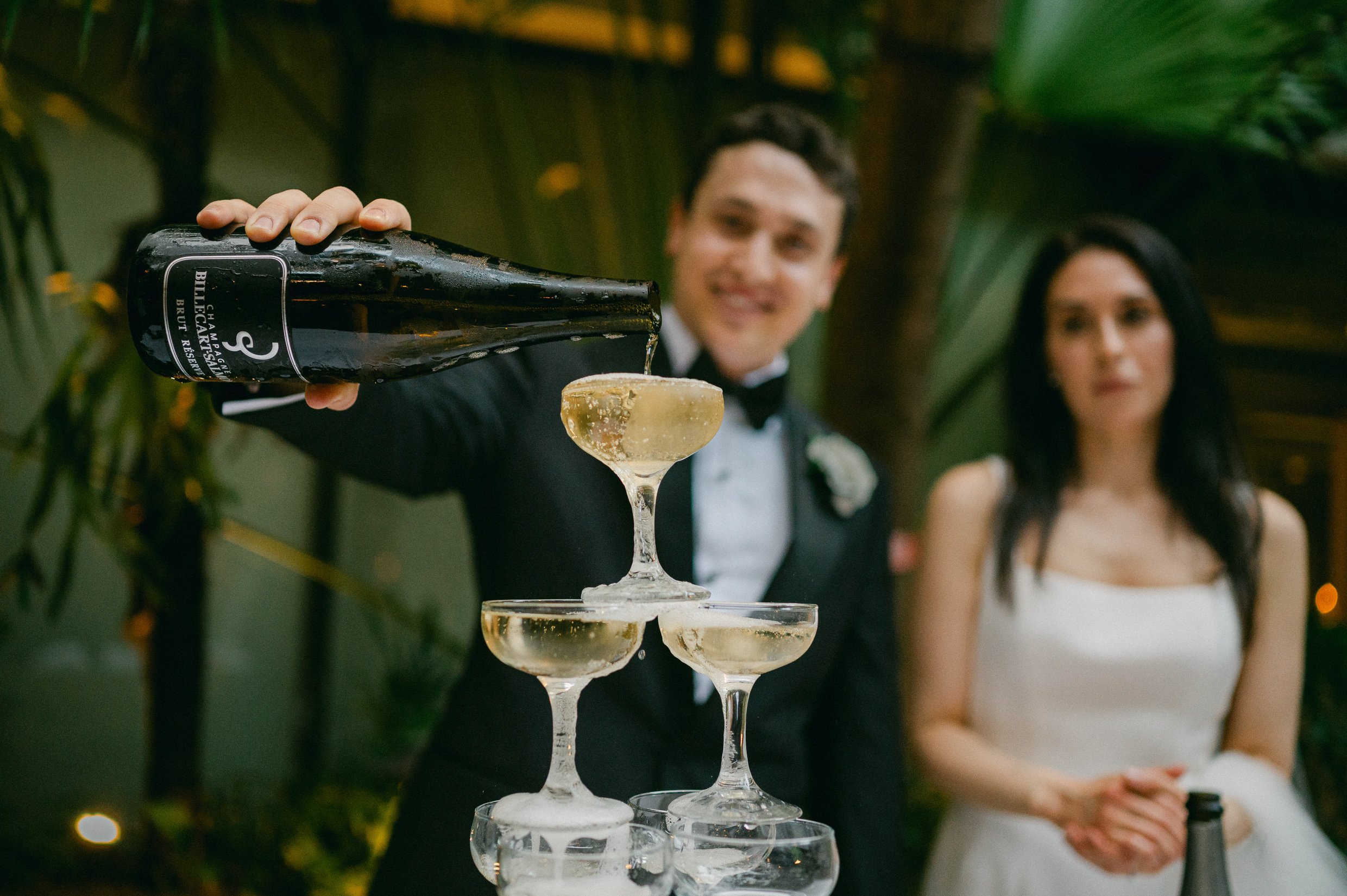 A groom pouring champagne into a tower of glasses, with his bride standing nearby at their Brennan's wedding reception in New Orleans.
