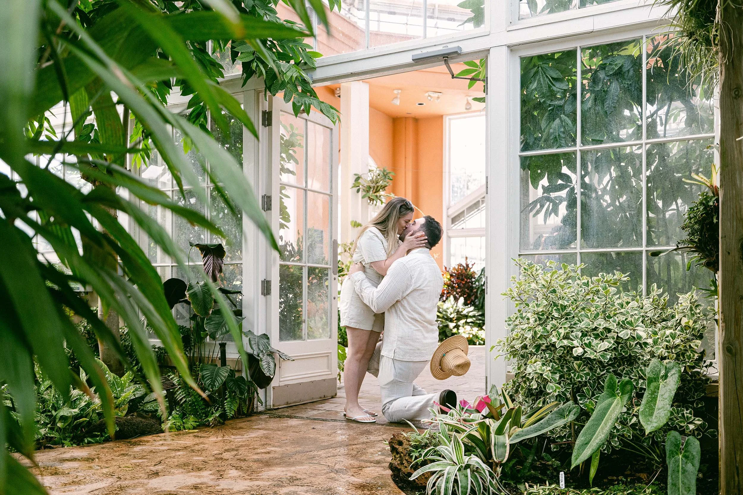 A man proposing marriage to a woman inside a lush greenhouse, surrounded by various plants and greenery, with the woman smiling and holding the man's face as he kneels.