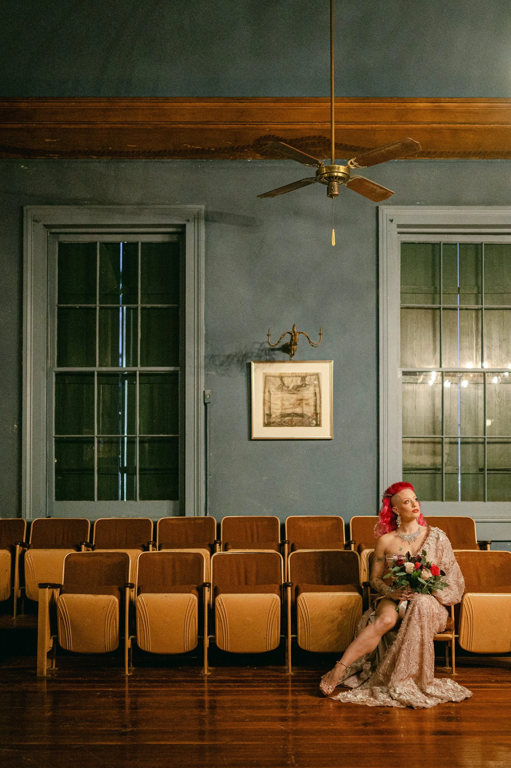 A portrait of a bride with bright pink hair sitting alone in an empty auditorium, wearing a pink lace gown and holding a bouquet of flowers.