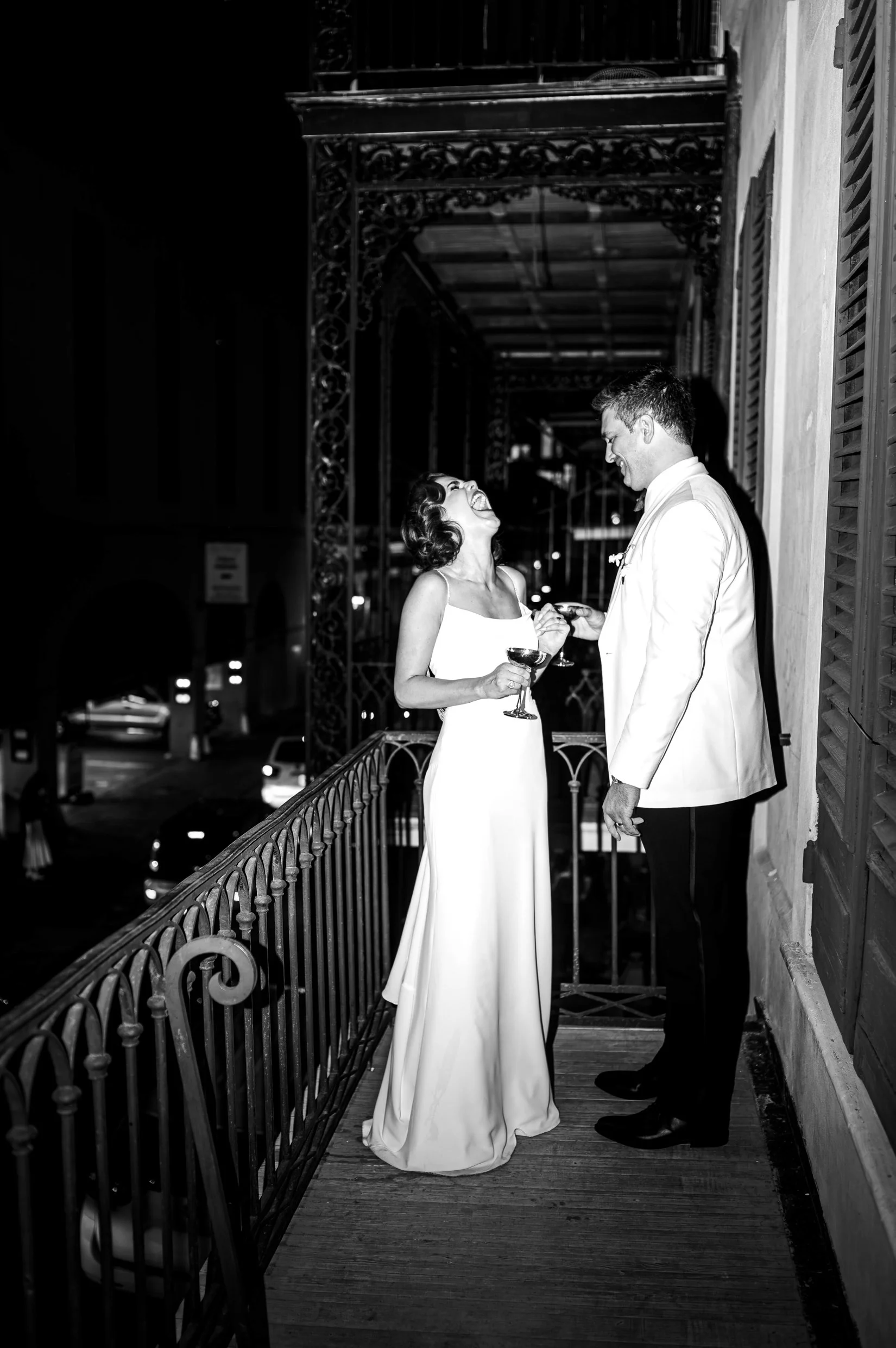 A black-and-white photo of a bride and groom standing on a balcony, laughing and holding hands, with New Orleans city lights in the background.
