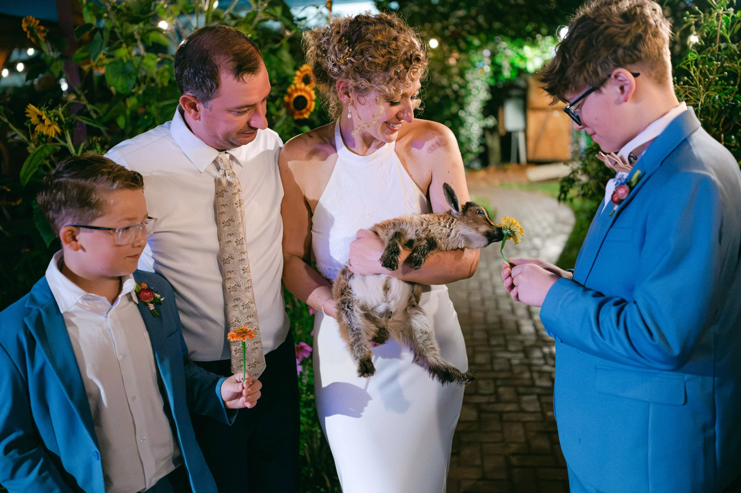 A wedding scene with a bride holding a baby goat and her groom and two sons. The background is decorated with green foliage and flowers.
