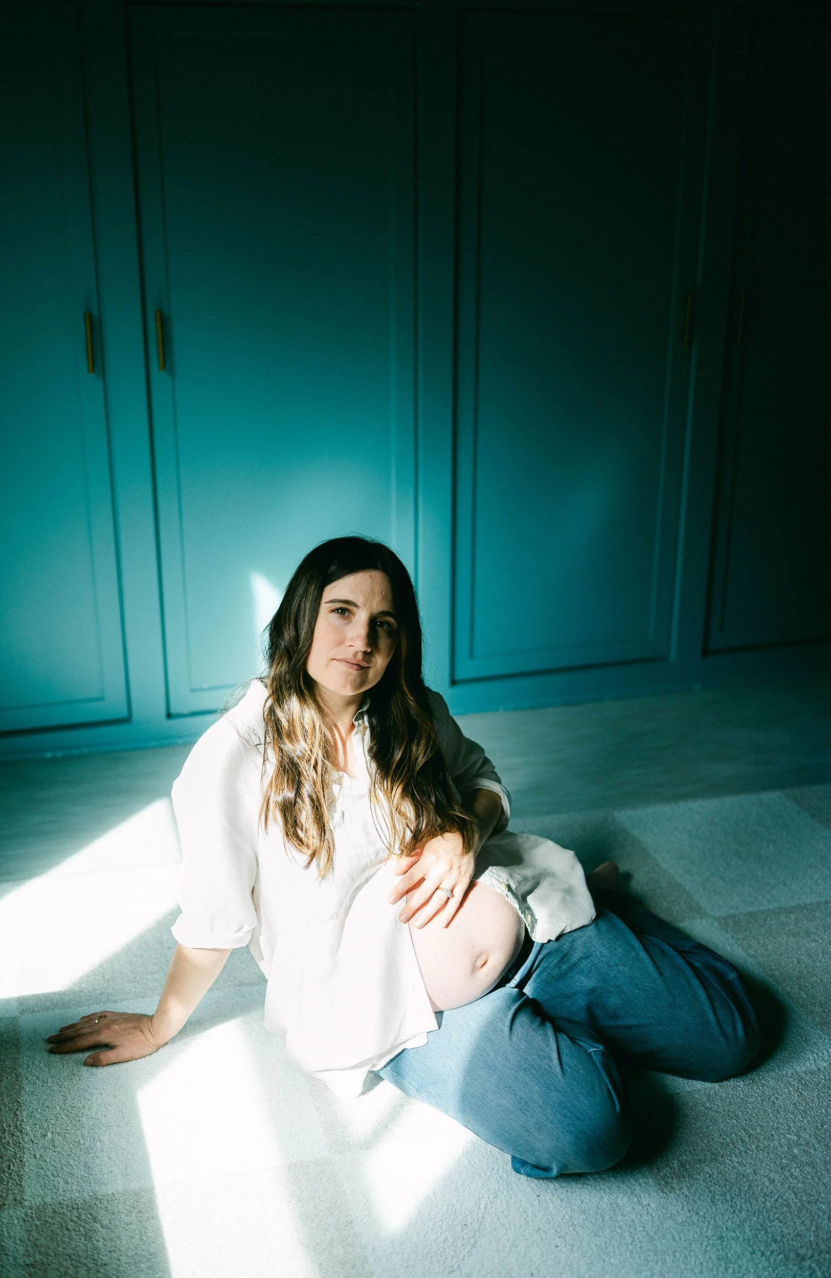 Pregnant woman with long brown hair sitting on the floor in a sunlit room with teal walls, wearing a white shirt and dark jeans.