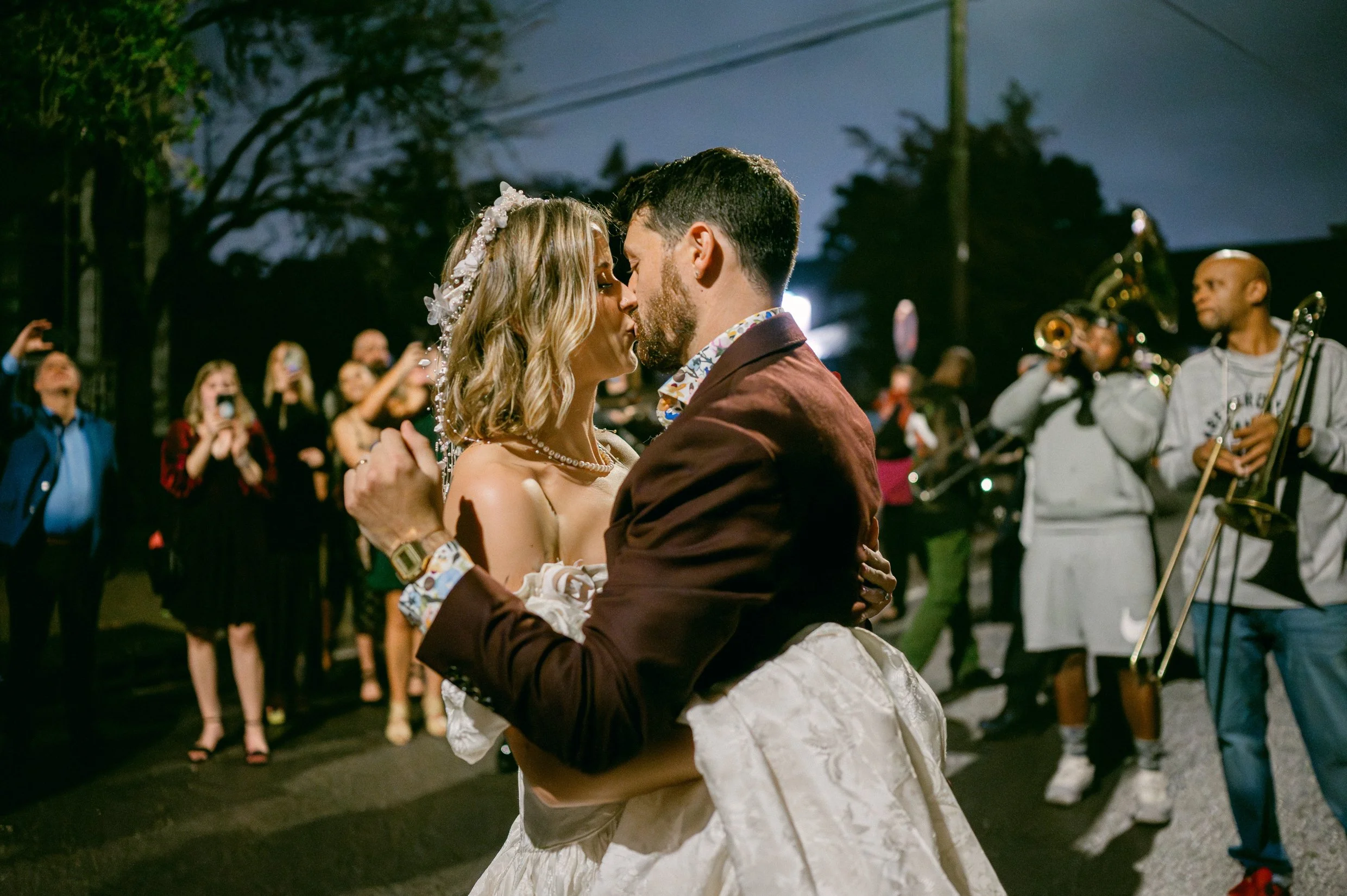 A bride and groom dancing closely at night, surrounded by onlookers and a live band playing instruments, with trees and streetlights in the background.