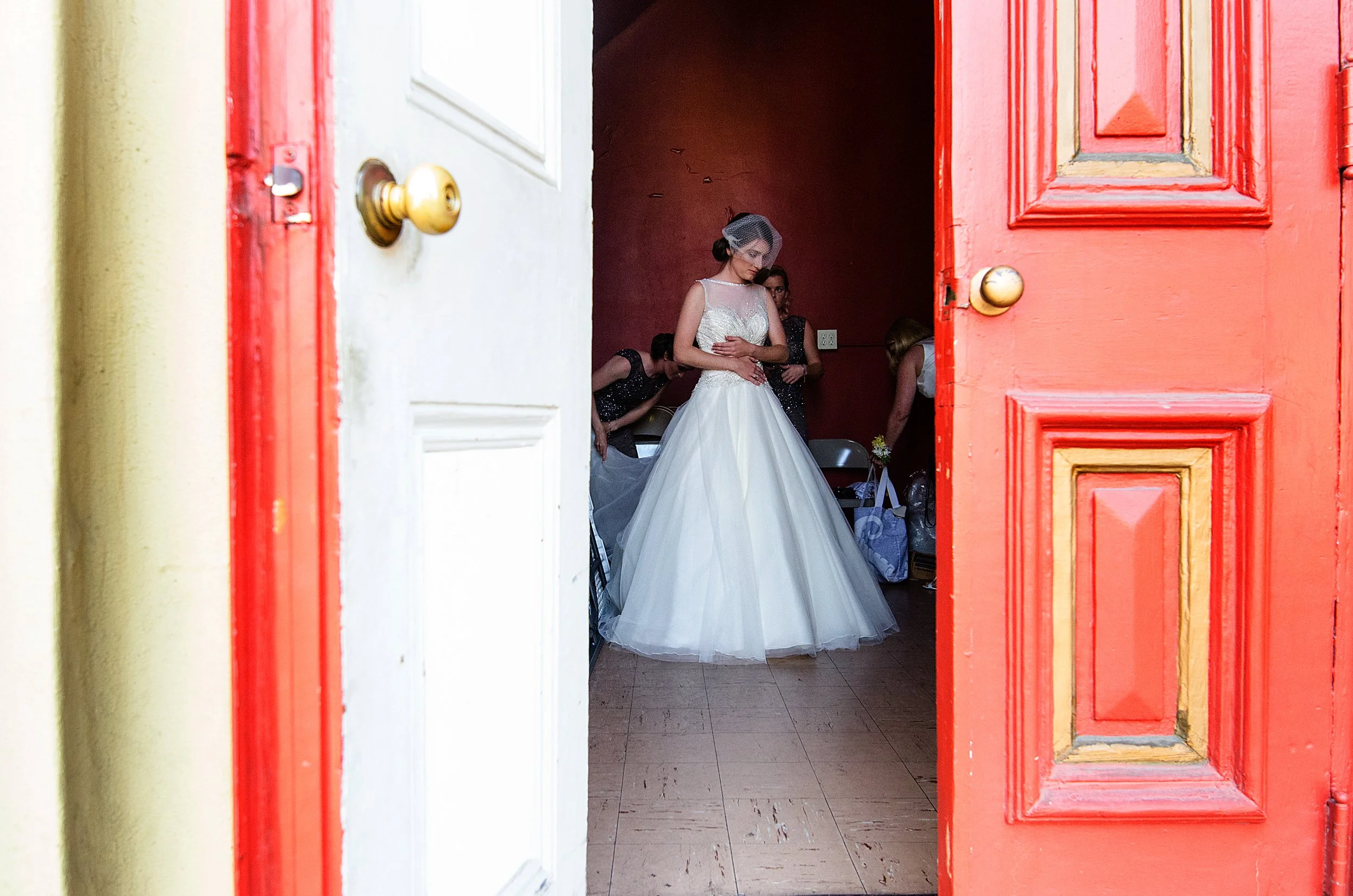 A bride in a wedding dress and veil standing inside a room, viewed through an open door.