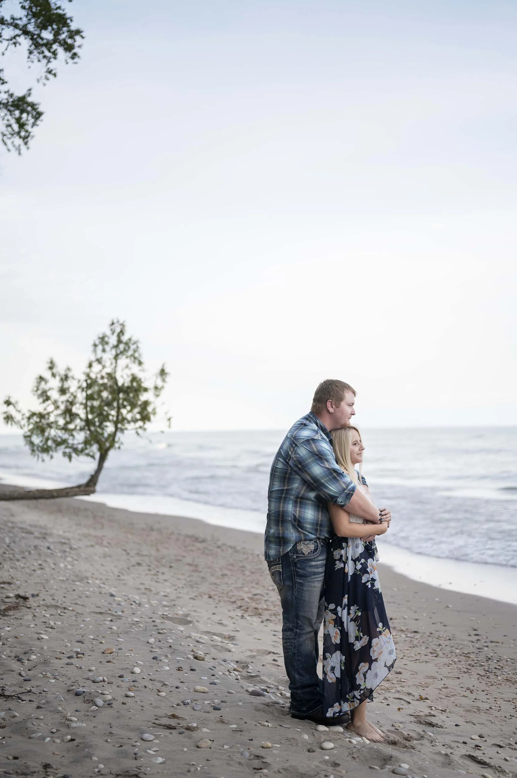 An engaged couple standing on a beach, embracing each other, with trees and the ocean in the background.