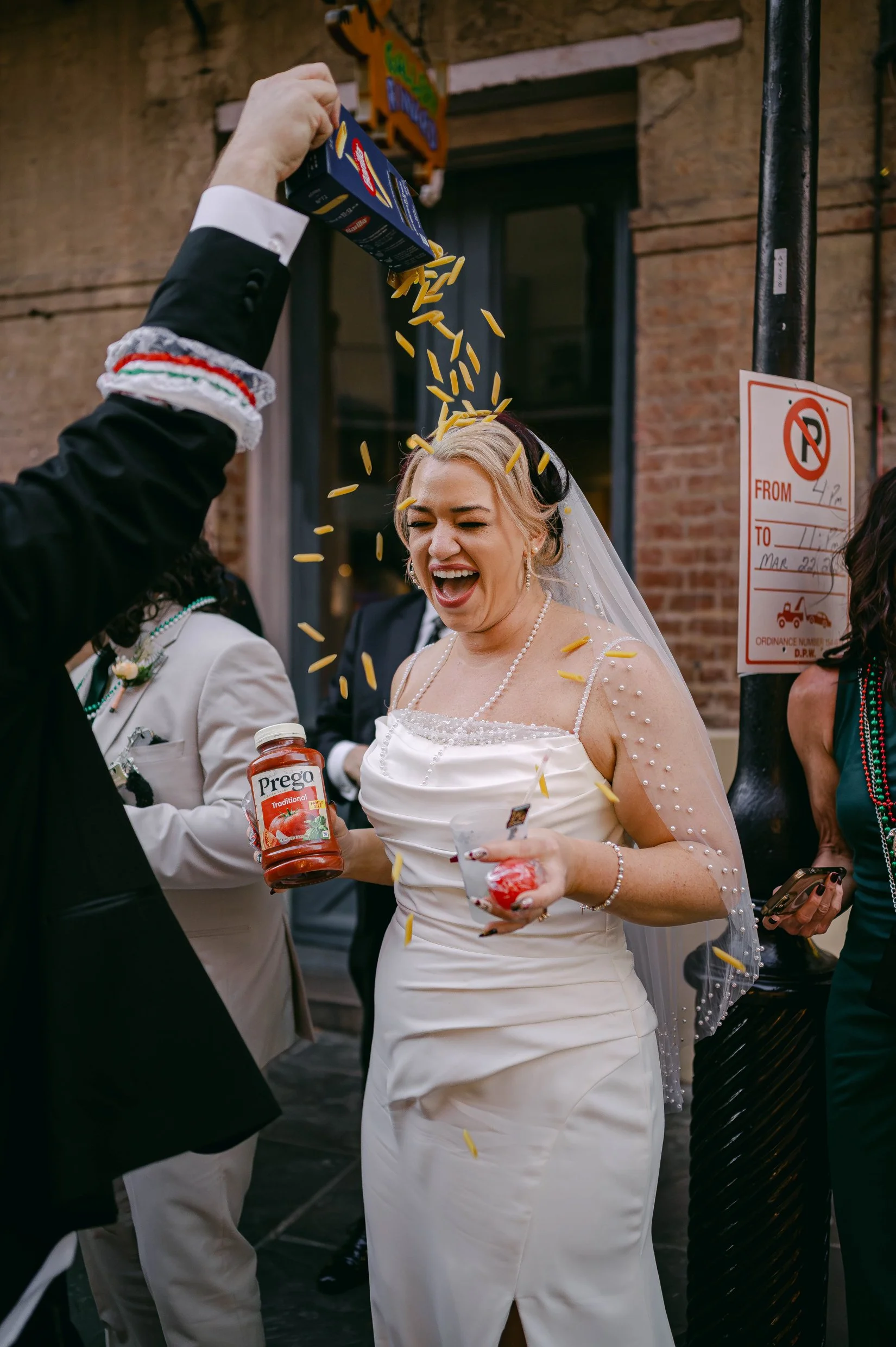 A bride in a white wedding dress uncook yellow pasta is poured over her head. She holds a jar of pasta sauce in one hand and a small container of red sauce in the other while watching the Italian parade in New Orleans.