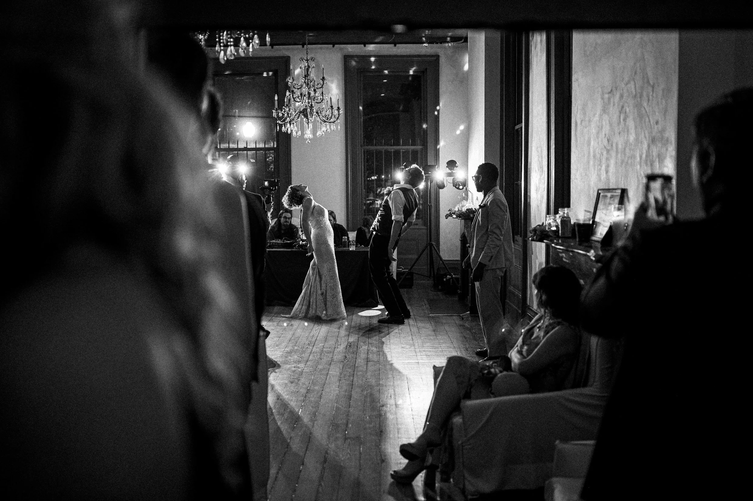 Black and white photo of a wedding reception with a bride and groom dancing, surrounded by guests, in an elegant room with large windows and a chandelier.