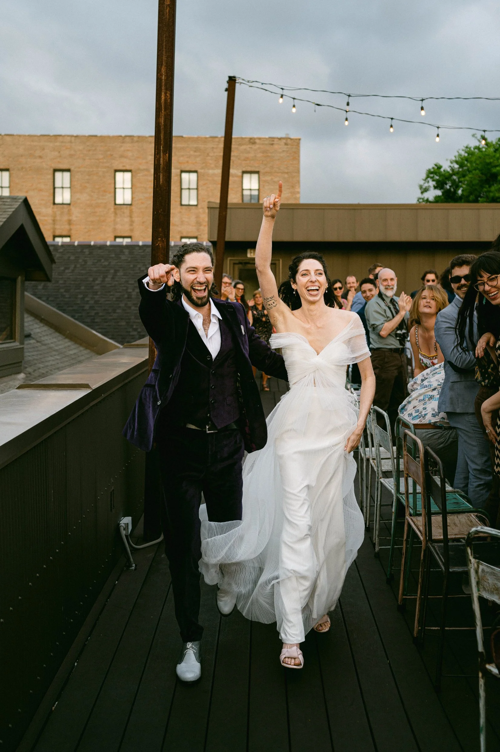 A bride and groom celebrating their wedding on a rooftop terrace, surrounded by friends and family, with outdoor string lights and city buildings in the background.