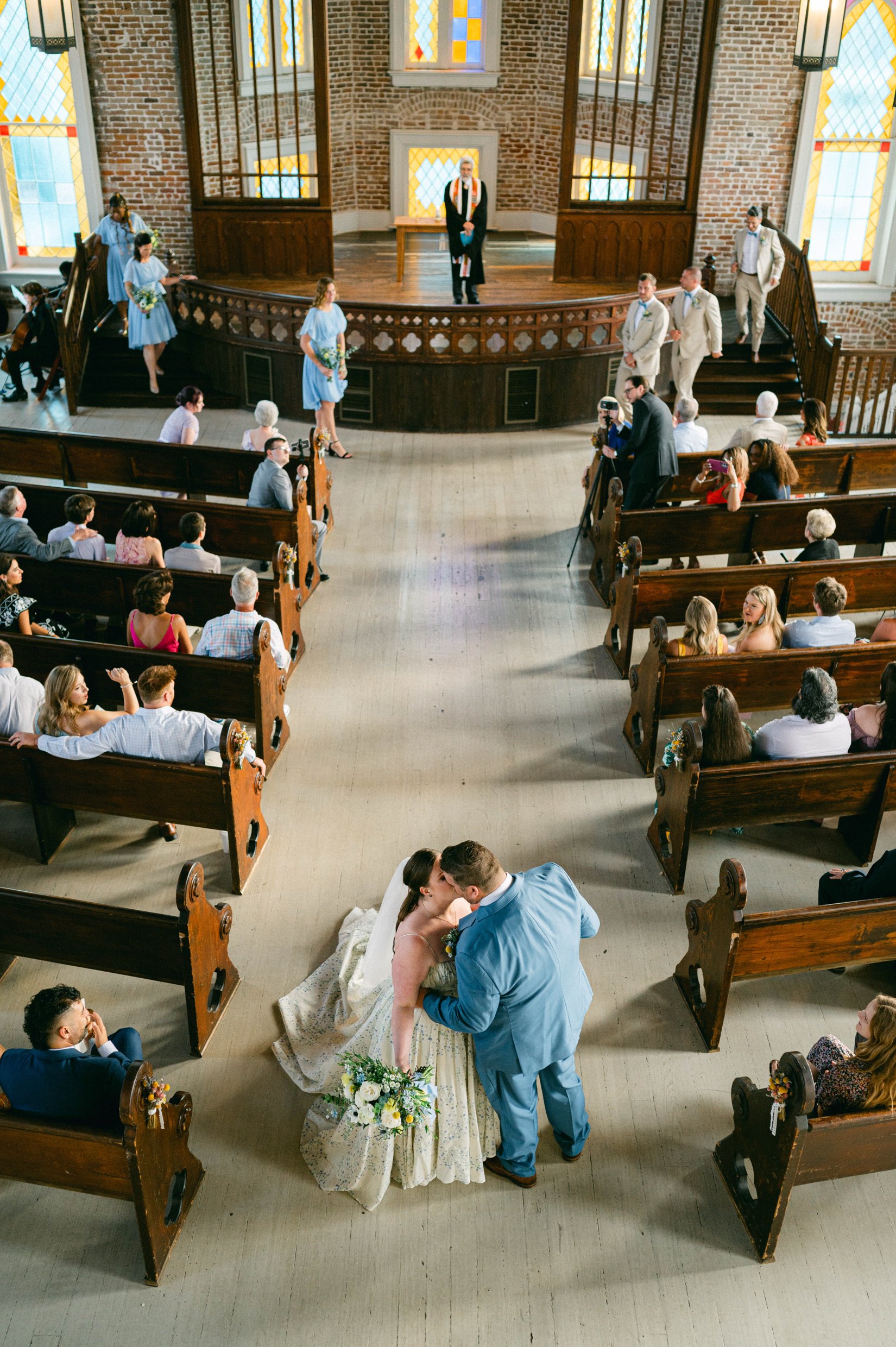 A wedding ceremony in a church with a bride and groom, officiant, and wedding party, with guests seated in pews and stained glass windows in the background.