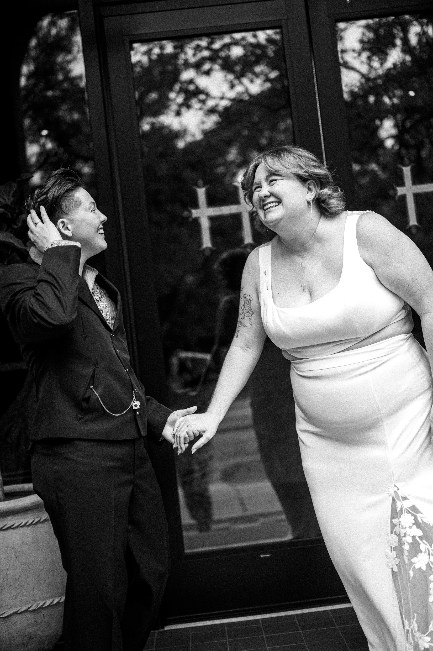 A queer couple are holding hands and smiling at each other. One spouse is wearing a suit and the other is in a white dress with floral embroidery. 