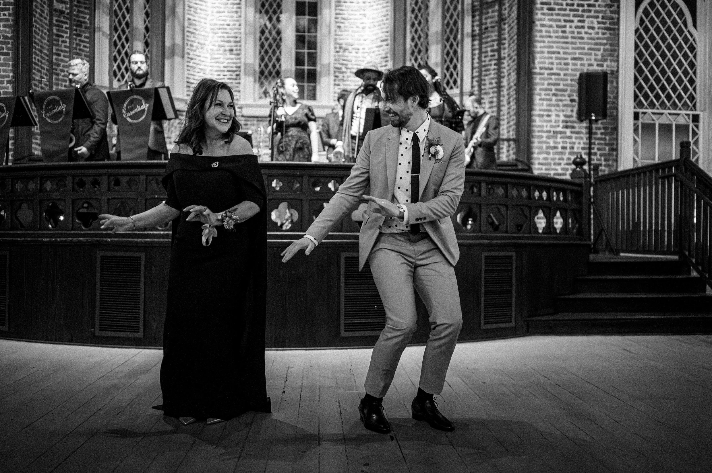 A groom and his mom dancing in a formal setting with a band playing in the background, all in black and white.