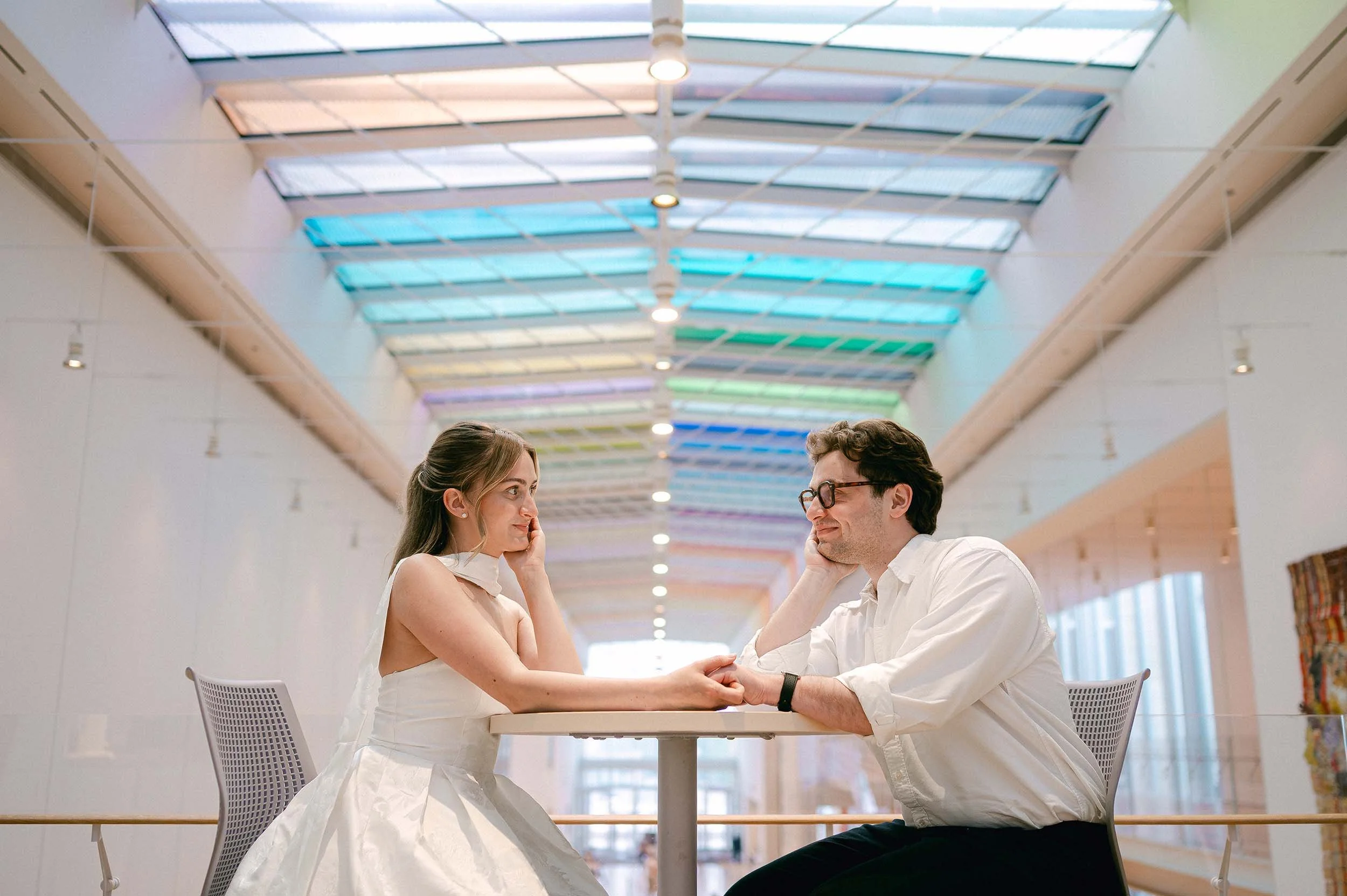 An engaged couple sitting across from each other at a table in a brightly lit, rainbow interior, indoor space, holding hands and facing each other smiling.