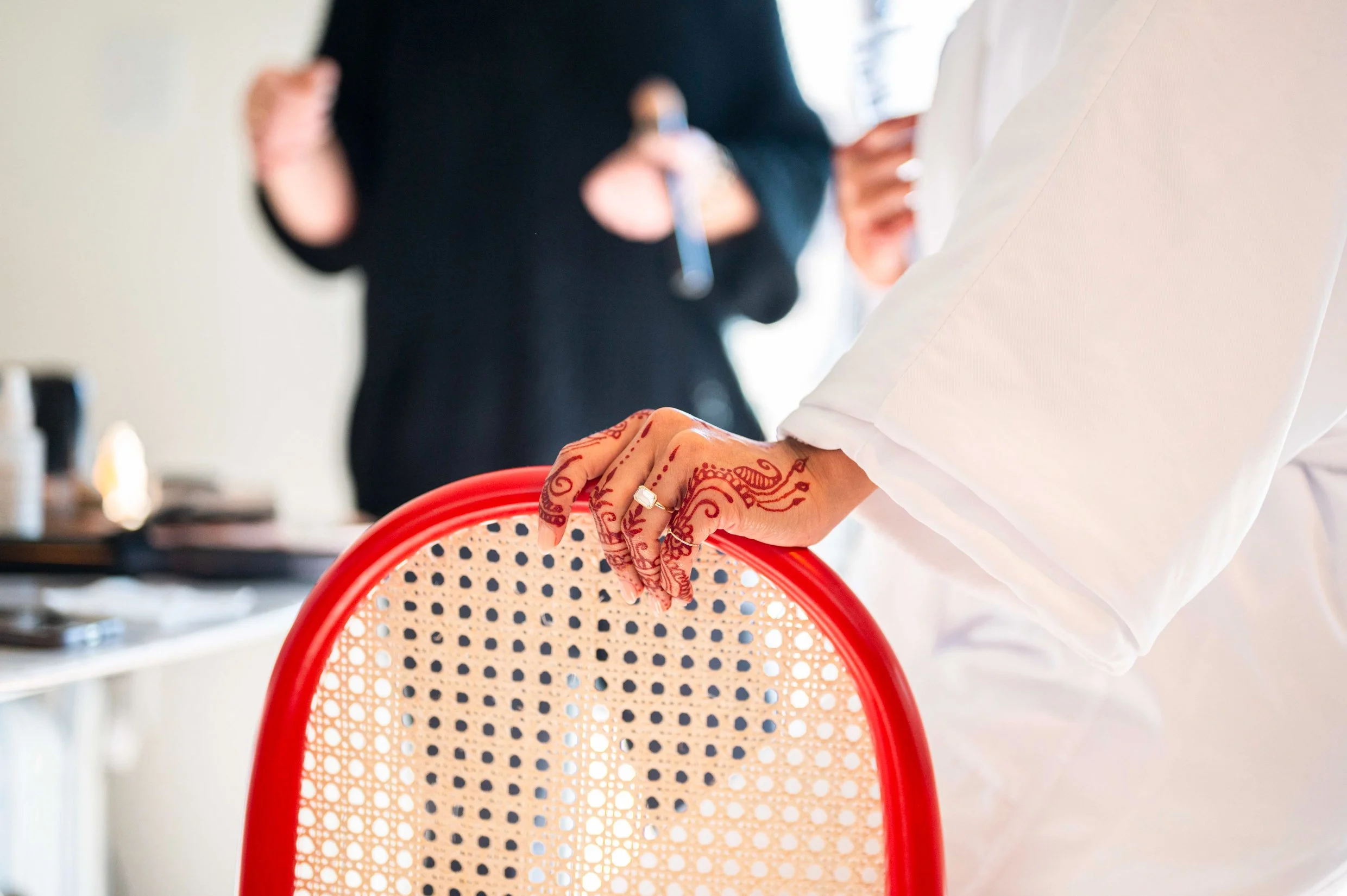 Close-up of a bride's hand with engagement ring and wedding henna designs, holding a red chair with a cane backrest.